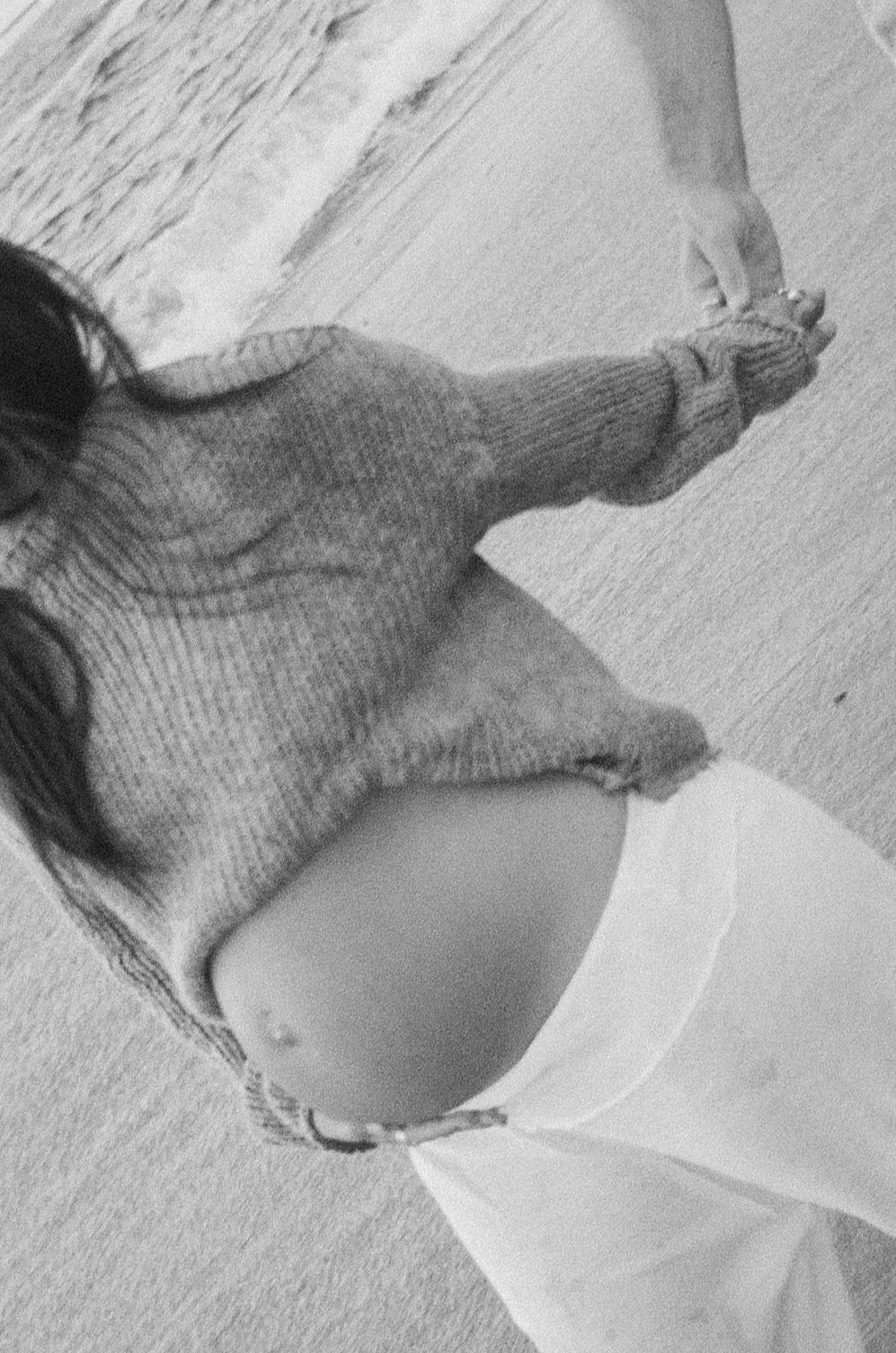 Black and white photo of a pregnant woman on a beach wearing a knit sweater and white pants.