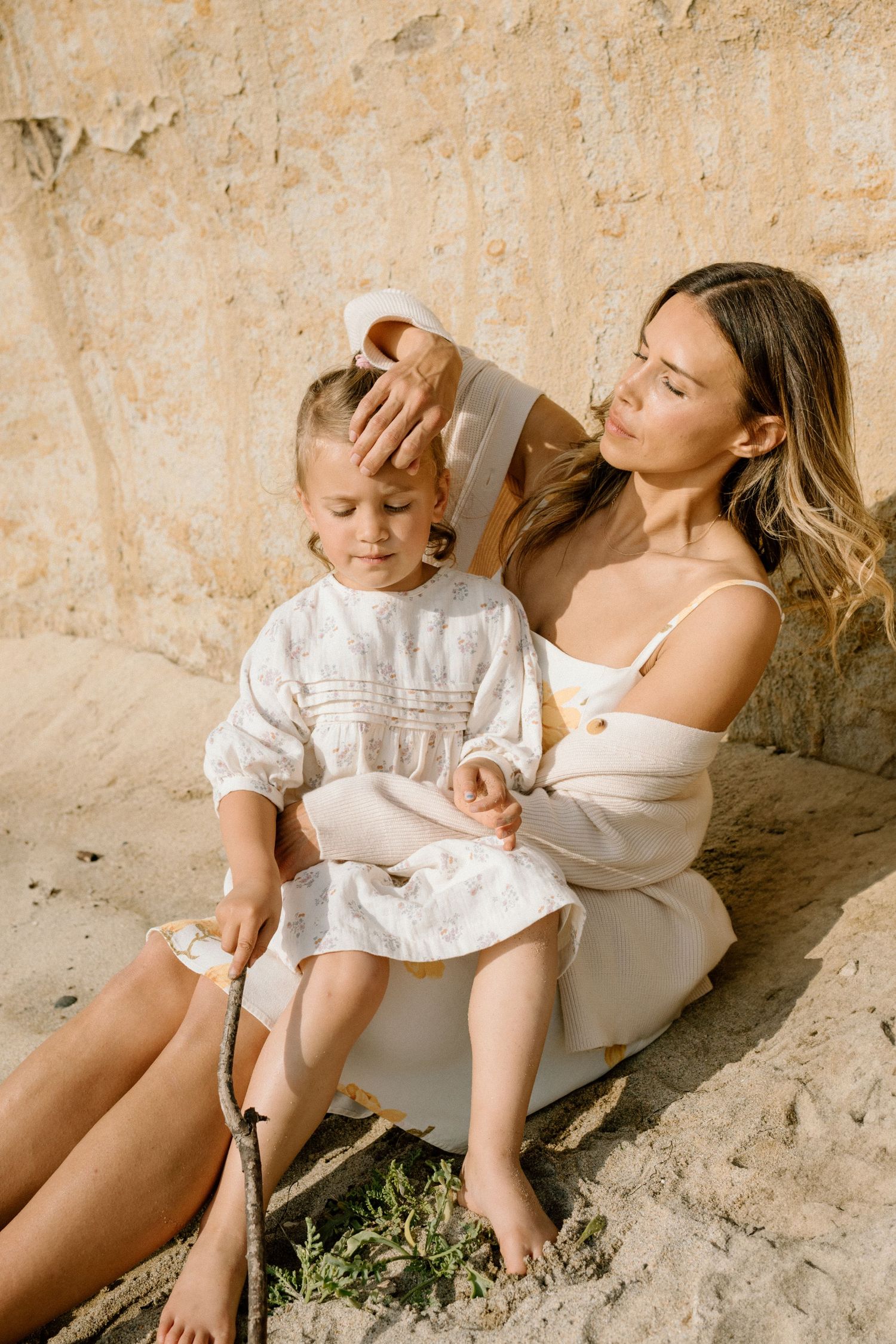 A mother tenderly fixes her young daughter's hair while sitting together on sandy ground in matching white outfits.