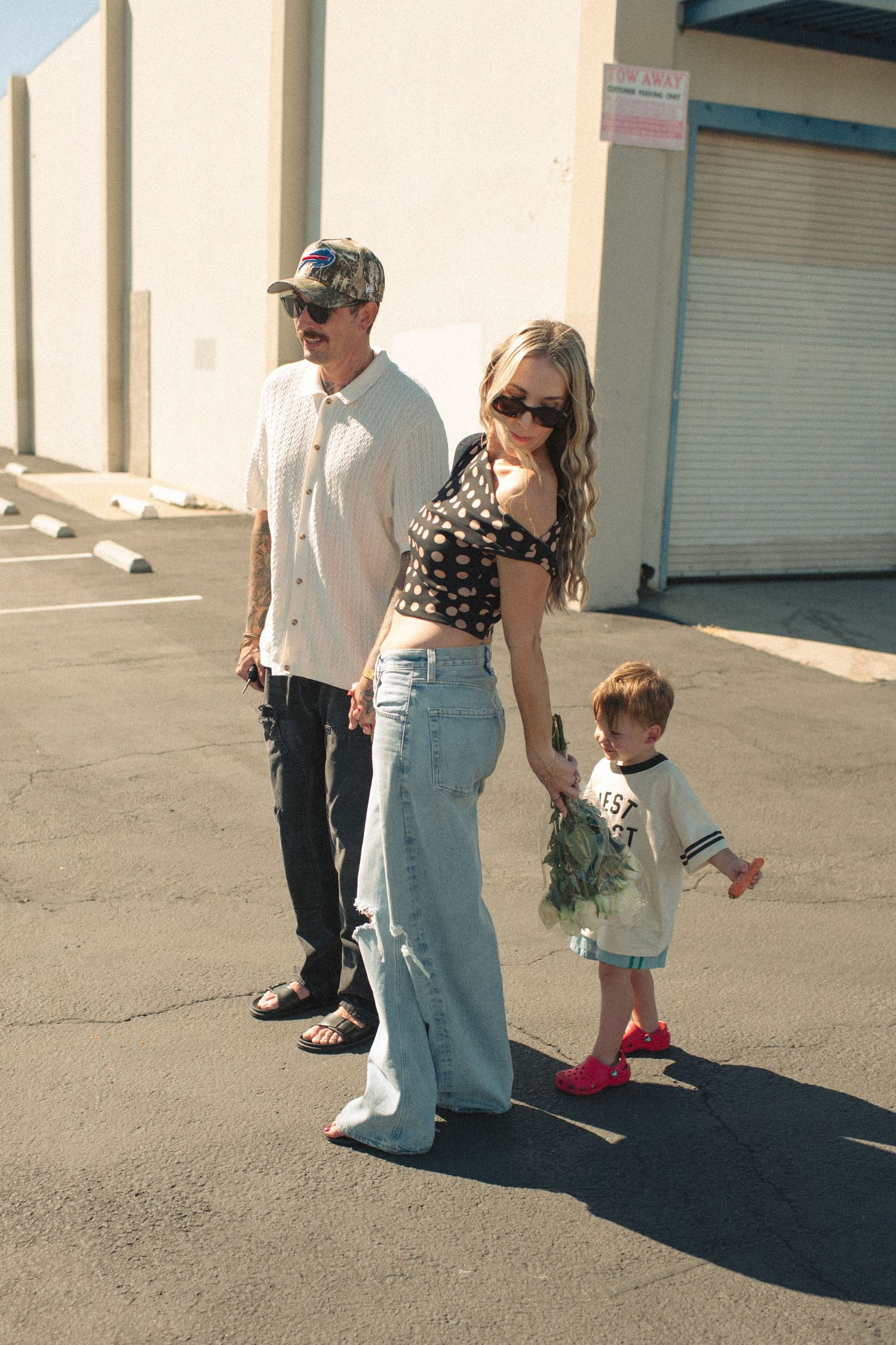 Young family with toddler walking through sunny parking lot, woman in crop top and wide-leg jeans, man in camo hat.
