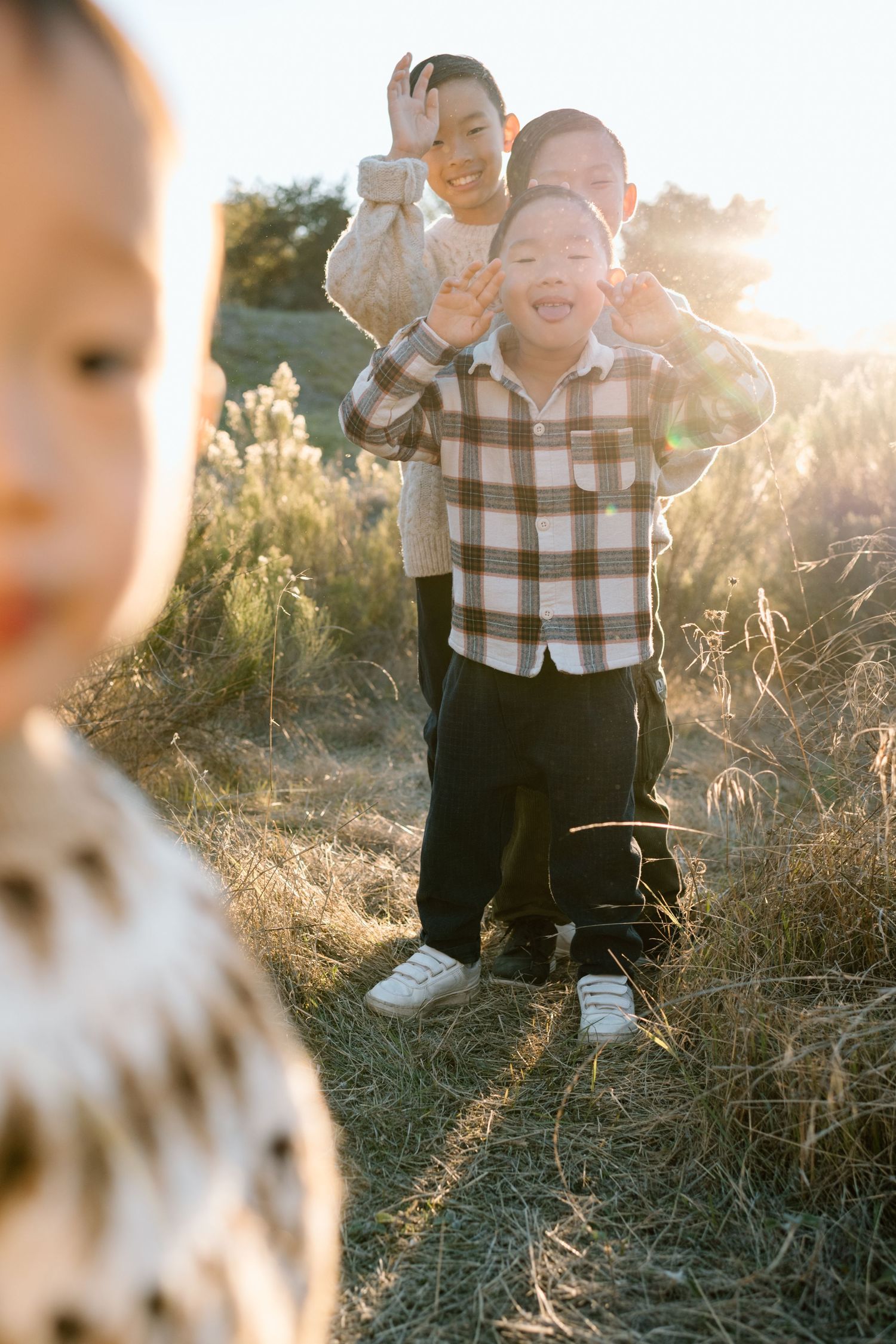 Three playful Asian boys making funny faces in a sunlit outdoor field at golden hour.