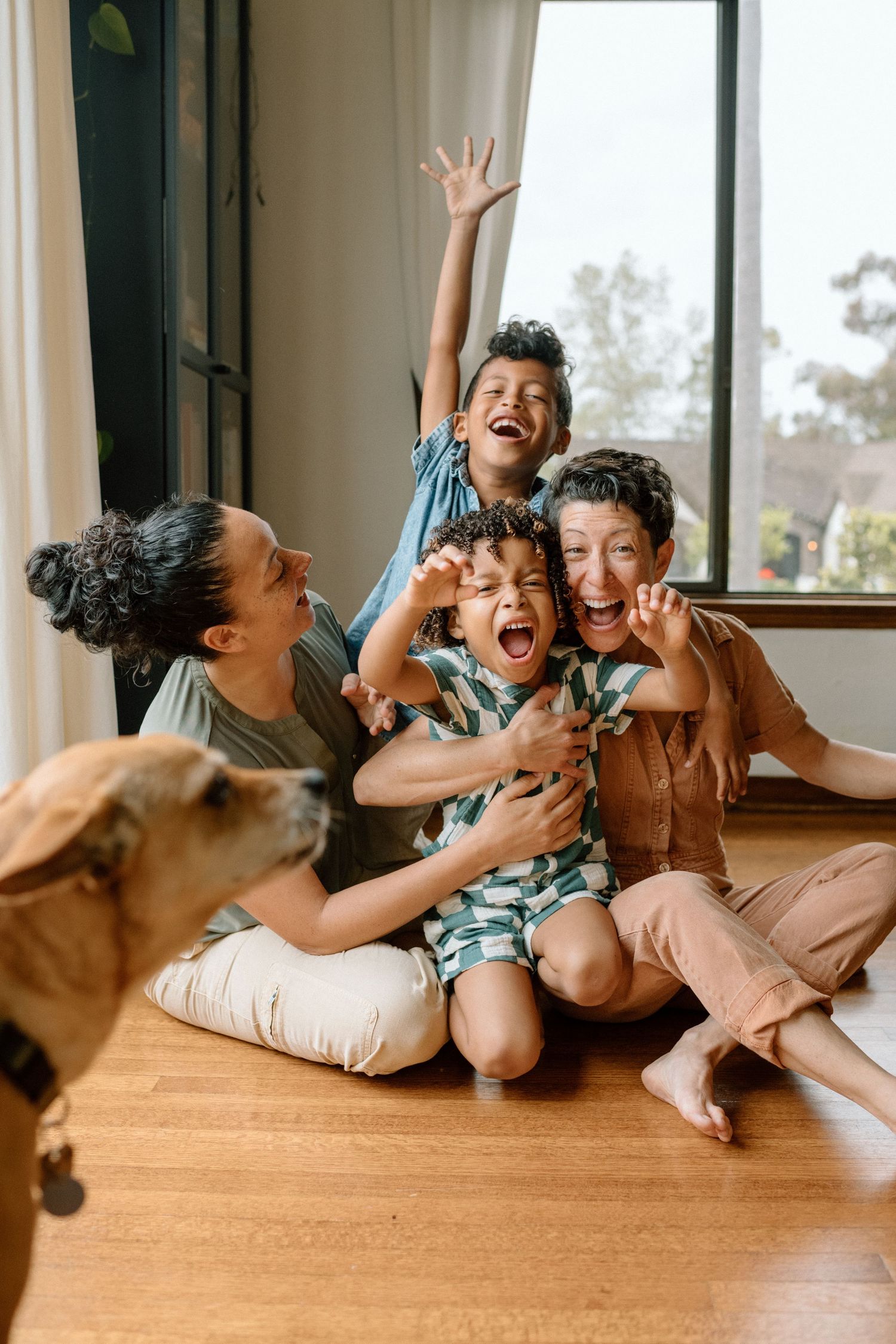 Joyful family with two moms, two kids, and a dog laughing together on hardwood floor.