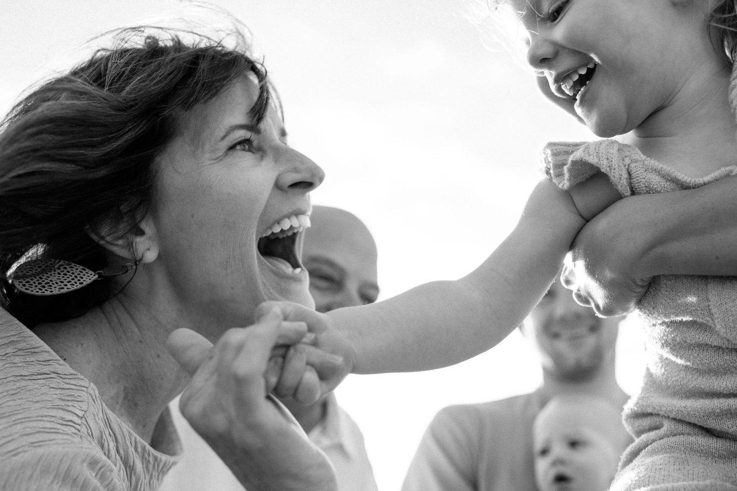 Black and white candid of laughing mother as toddler reaches for her face outdoors.