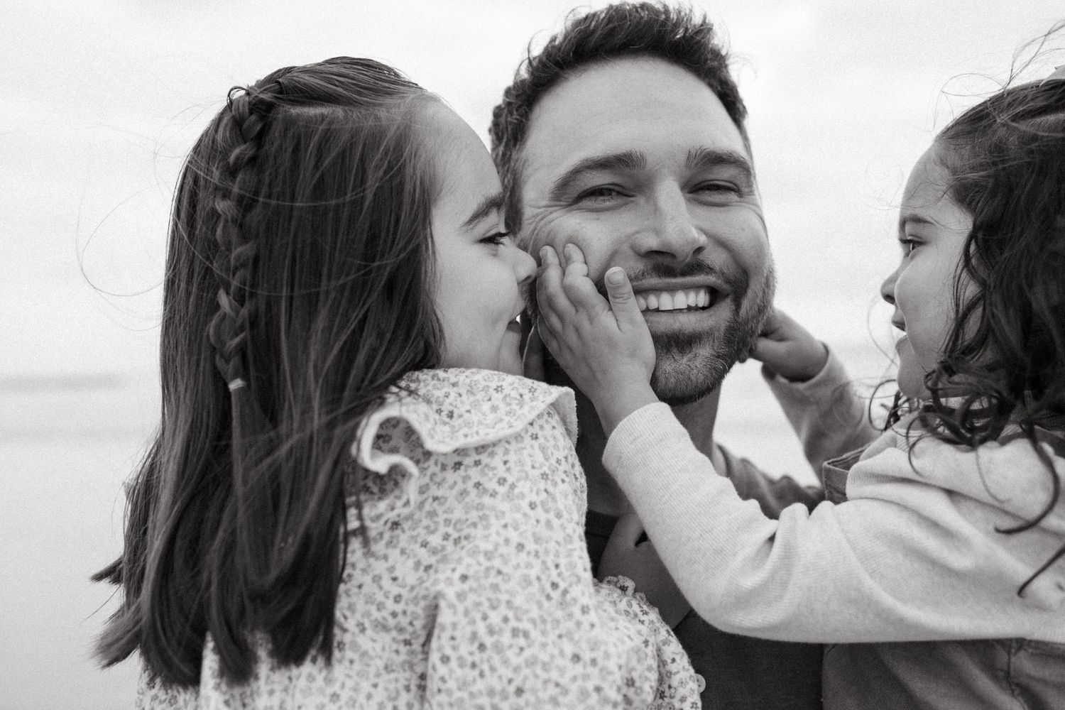 Black and white photo of a smiling man with two young girls kissing and touching his cheeks outdoors.