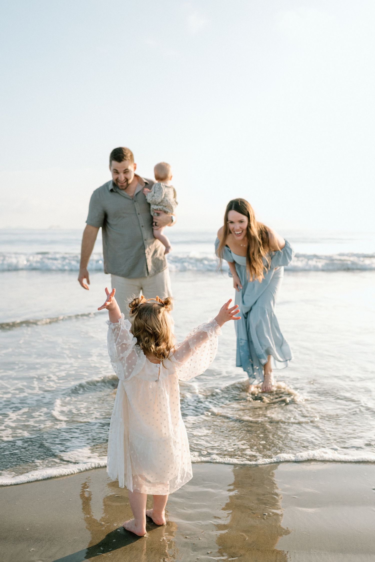 Young girl in white lace dress reaching toward parents at shoreline.