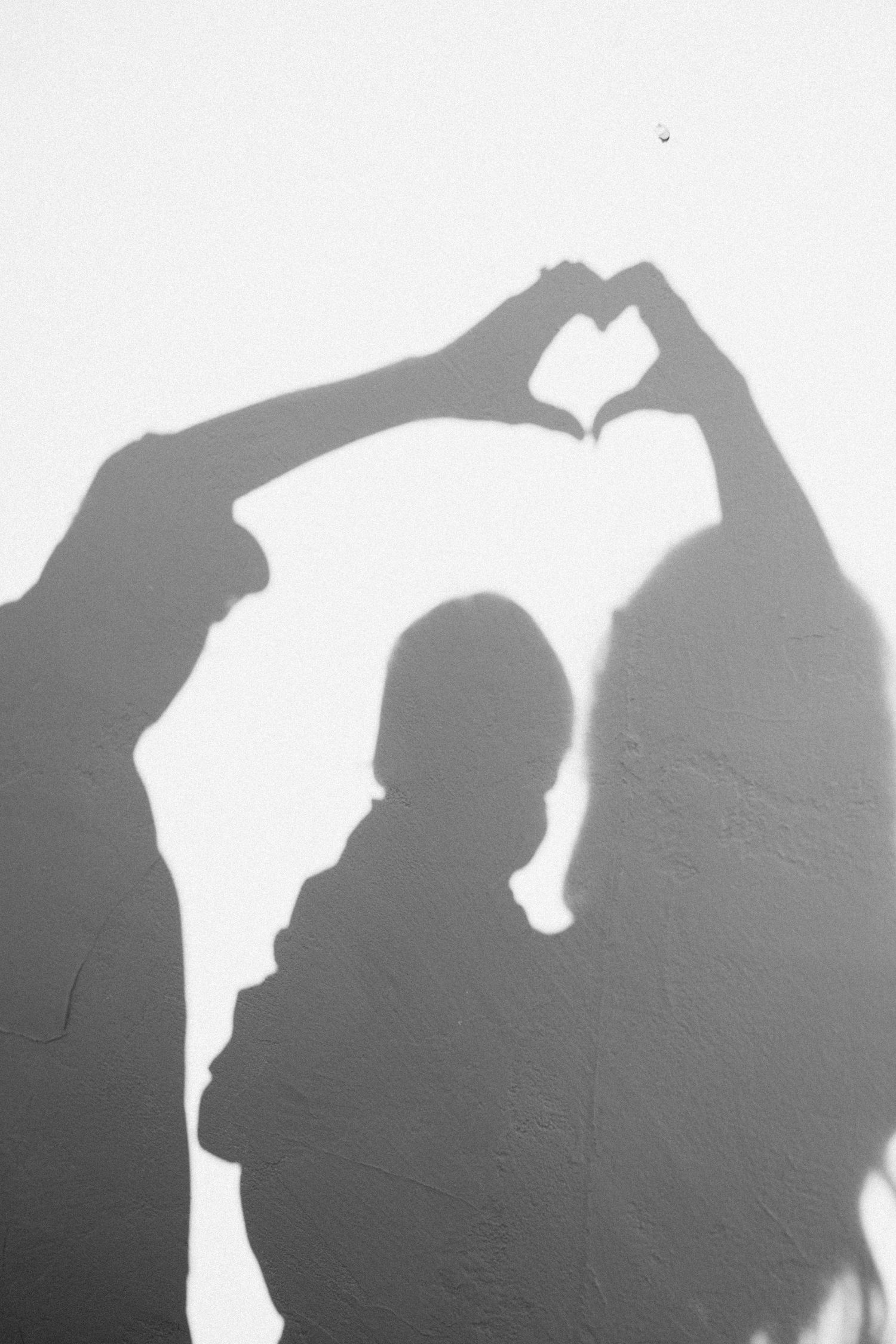 A shadow silhouette on a white wall shows two people making a heart shape with their hands against bright light.