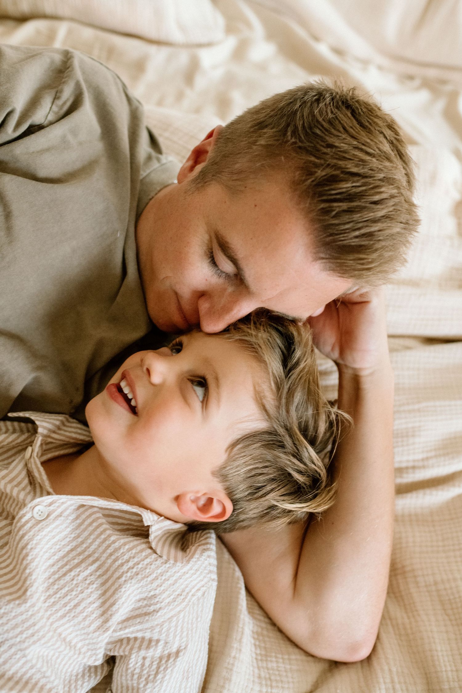 A father and young son share a tender moment lying together on a beige textured bed.