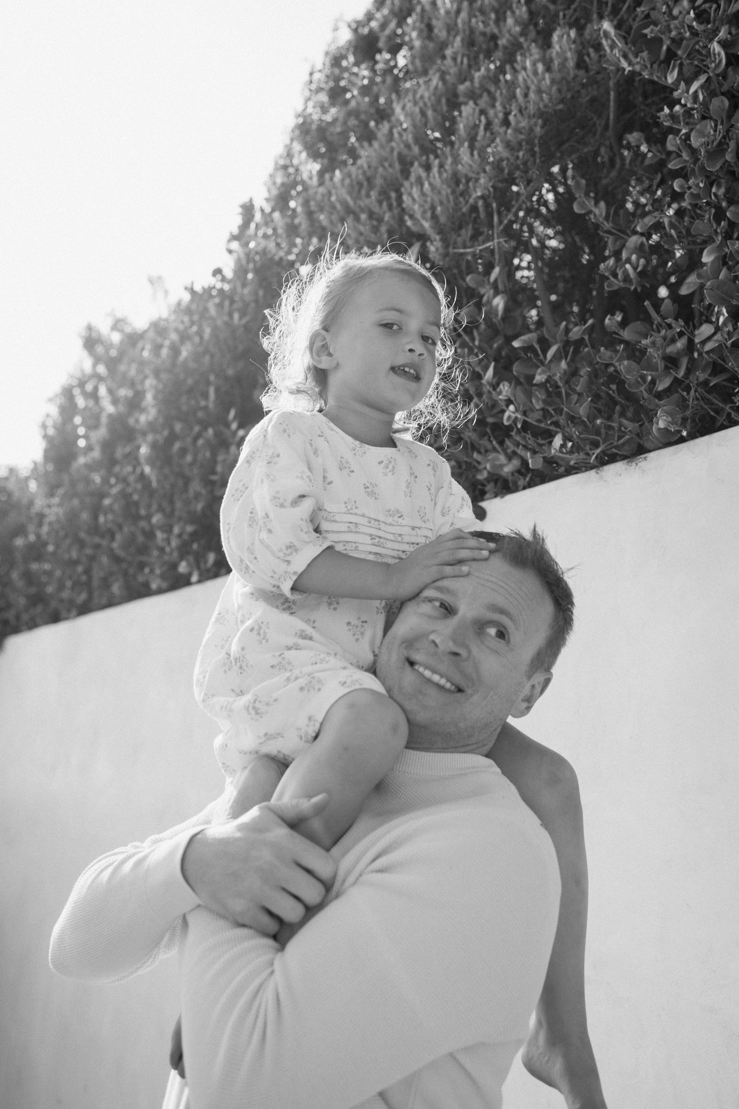 A smiling father carries his young daughter on his shoulders outdoors near a white wall with green hedges.