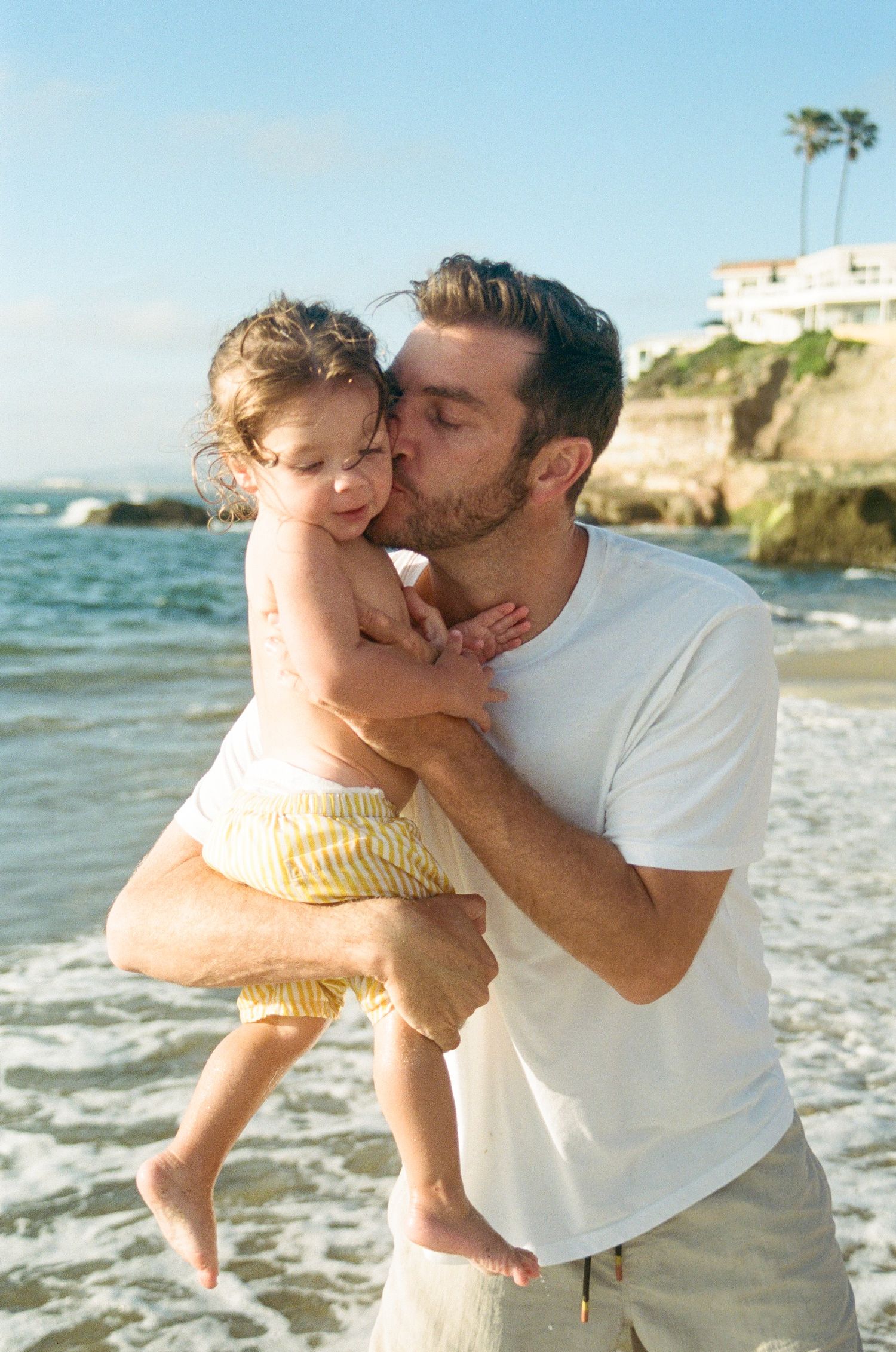 A father kisses his toddler on the cheek while holding them at a sunny beach with ocean waves.