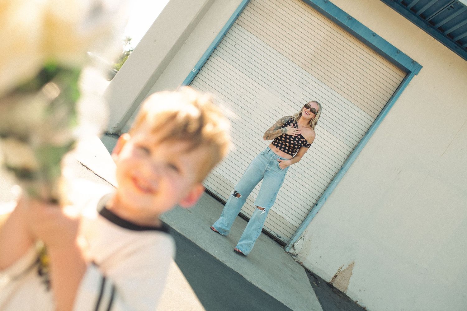 Blurred toddler in foreground with stylish mom in polka dot top and ripped jeans standing by garage door.
