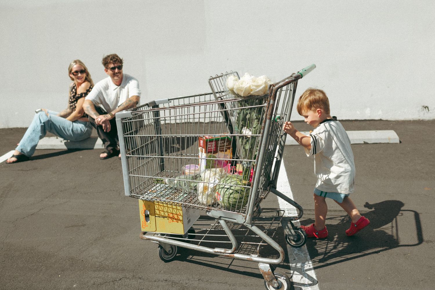 A young child pushes a full shopping cart in a parking lot while two adults sit relaxed against a wall in sunlight.