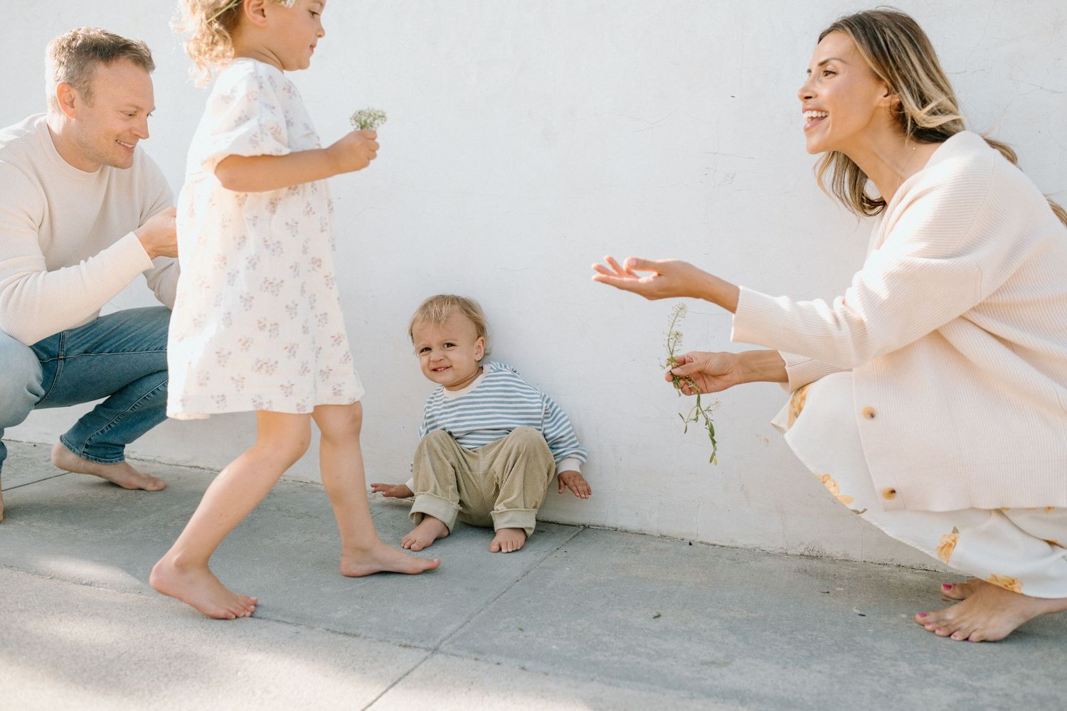 A joyful family with two young children playing outdoors against a white wall in soft natural light.