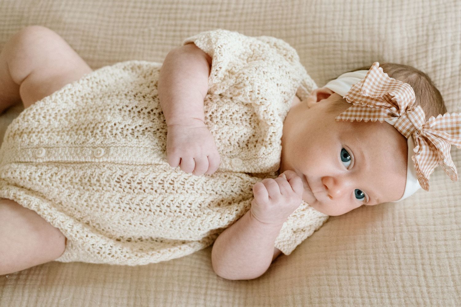 Newborn baby in cream knit romper with gingham bow headband lying on a beige blanket.