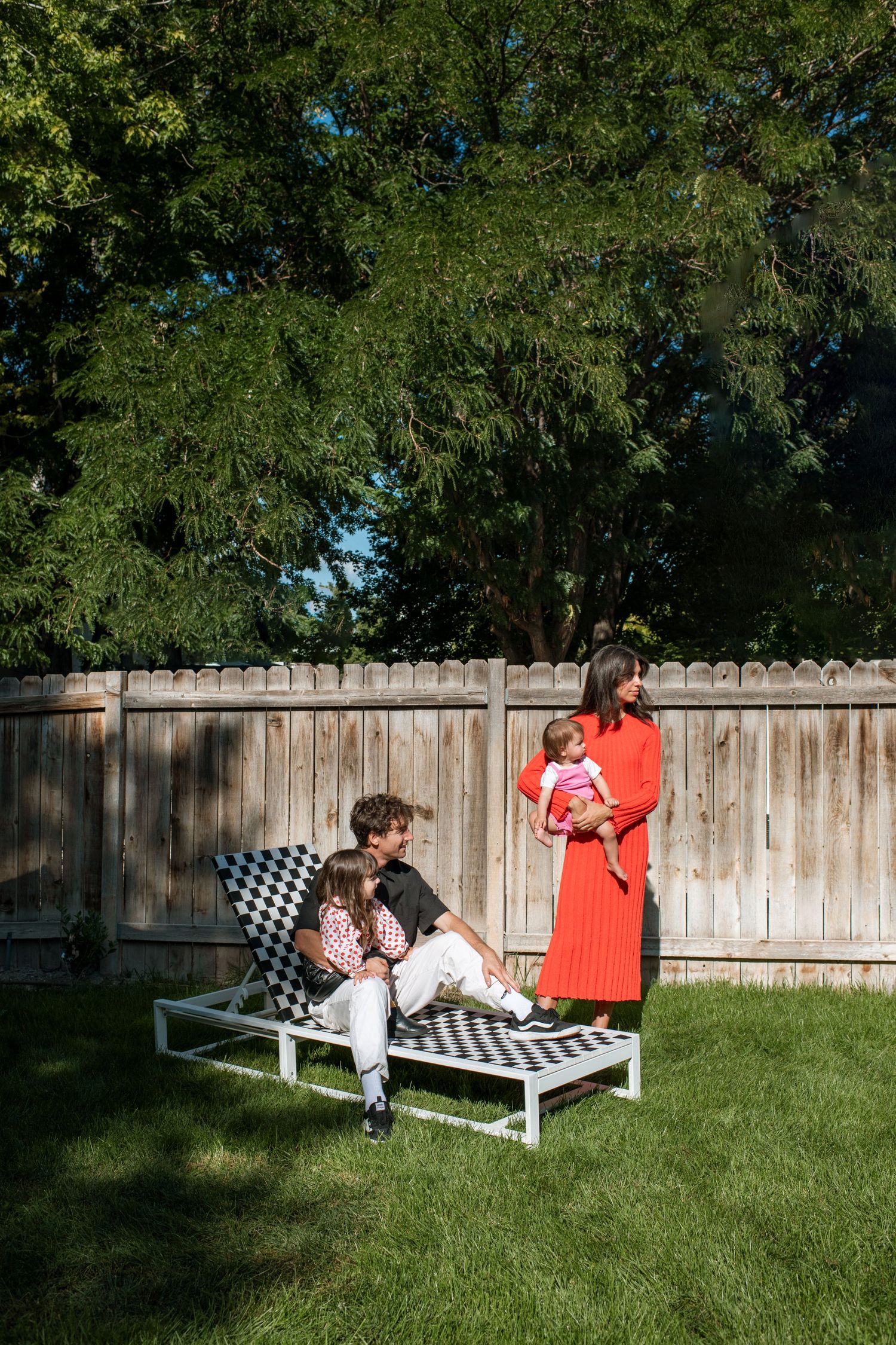 Family relaxing in sunny backyard, man and child on checkered lounge chair, woman in red dress holding baby near wooden fence.