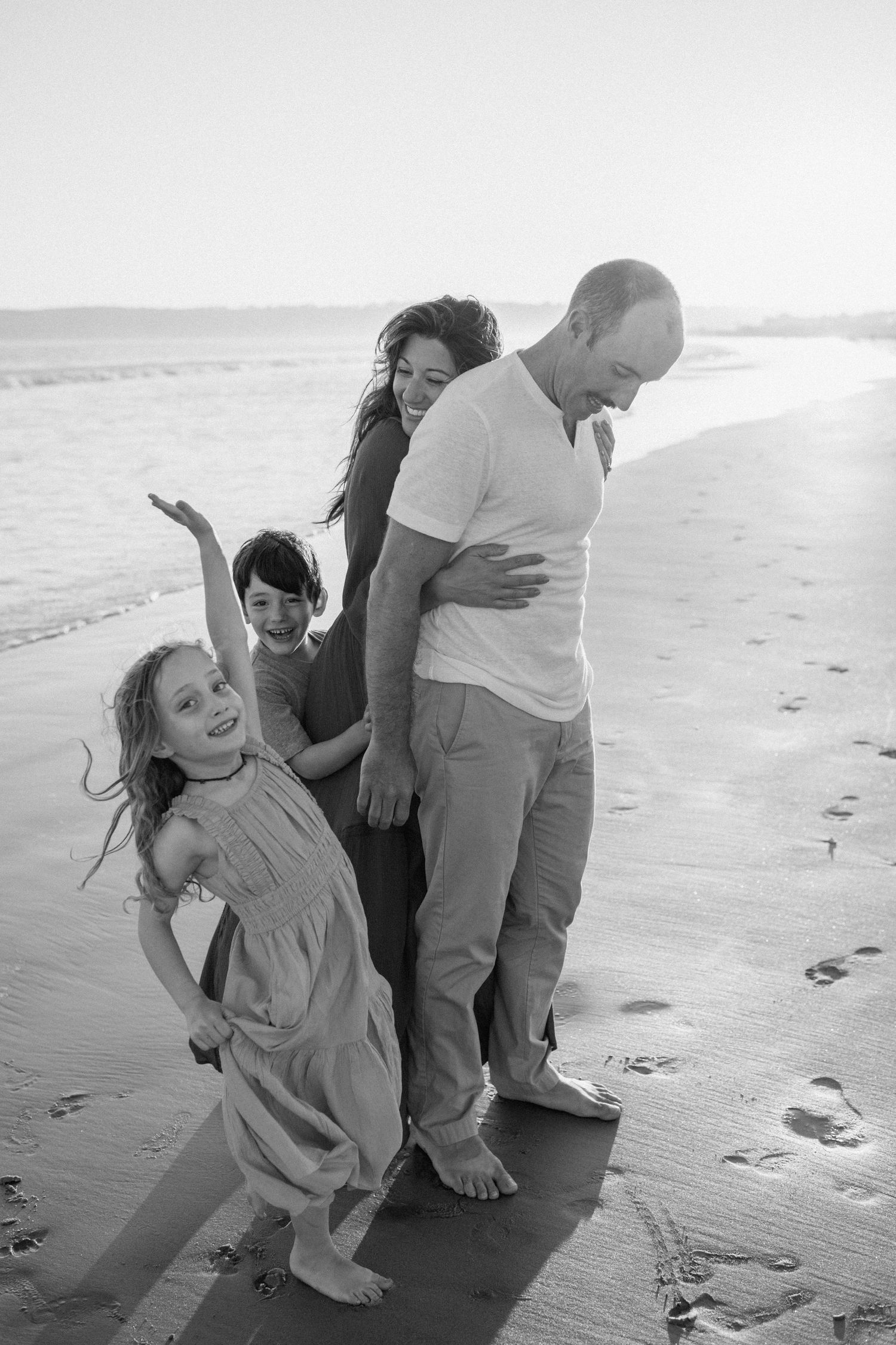 Black and white photo of a joyful family of four walking barefoot on a sunny beach.