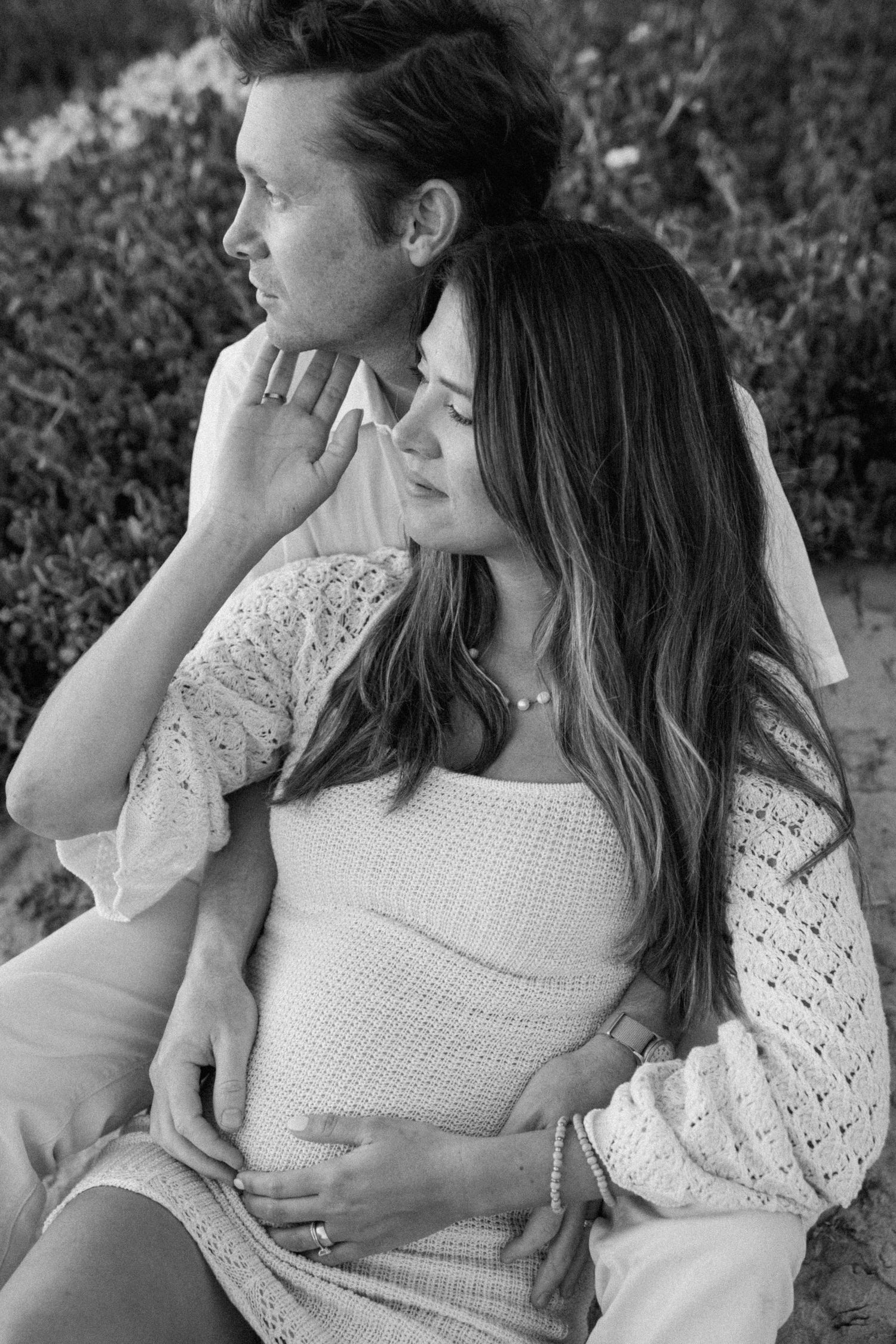 Black and white photo of a couple sitting together, man embracing pregnant woman outdoors.