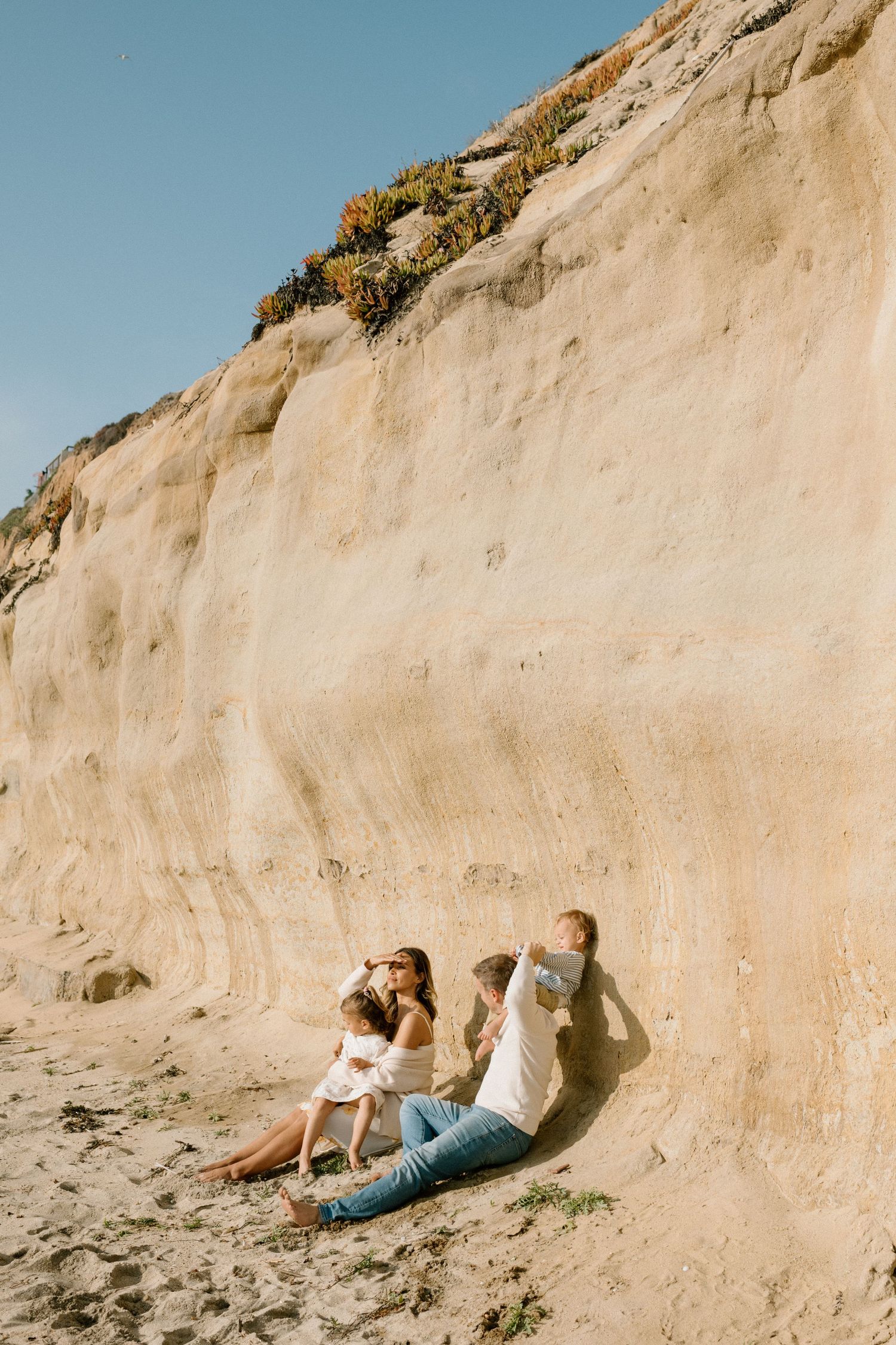 A family relaxes against a tall sandy cliff on a sunny beach, enjoying a peaceful outdoor moment together.