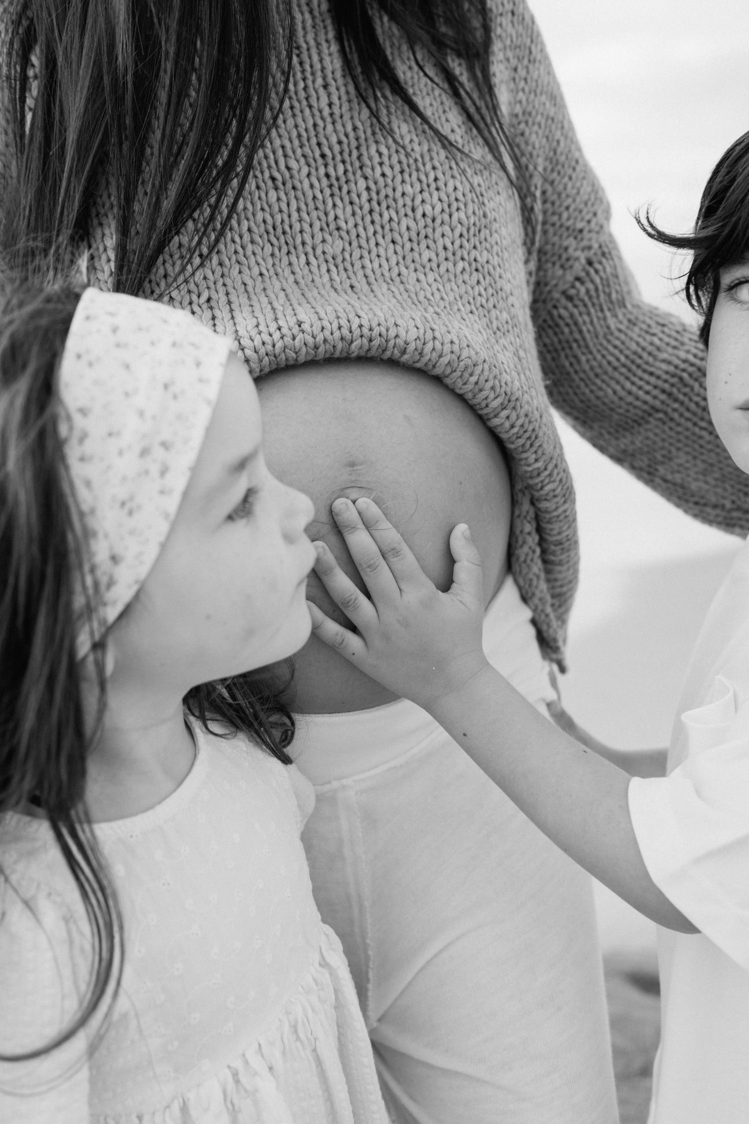 Young girl kissing pregnant mothers belly on beach in black and white.