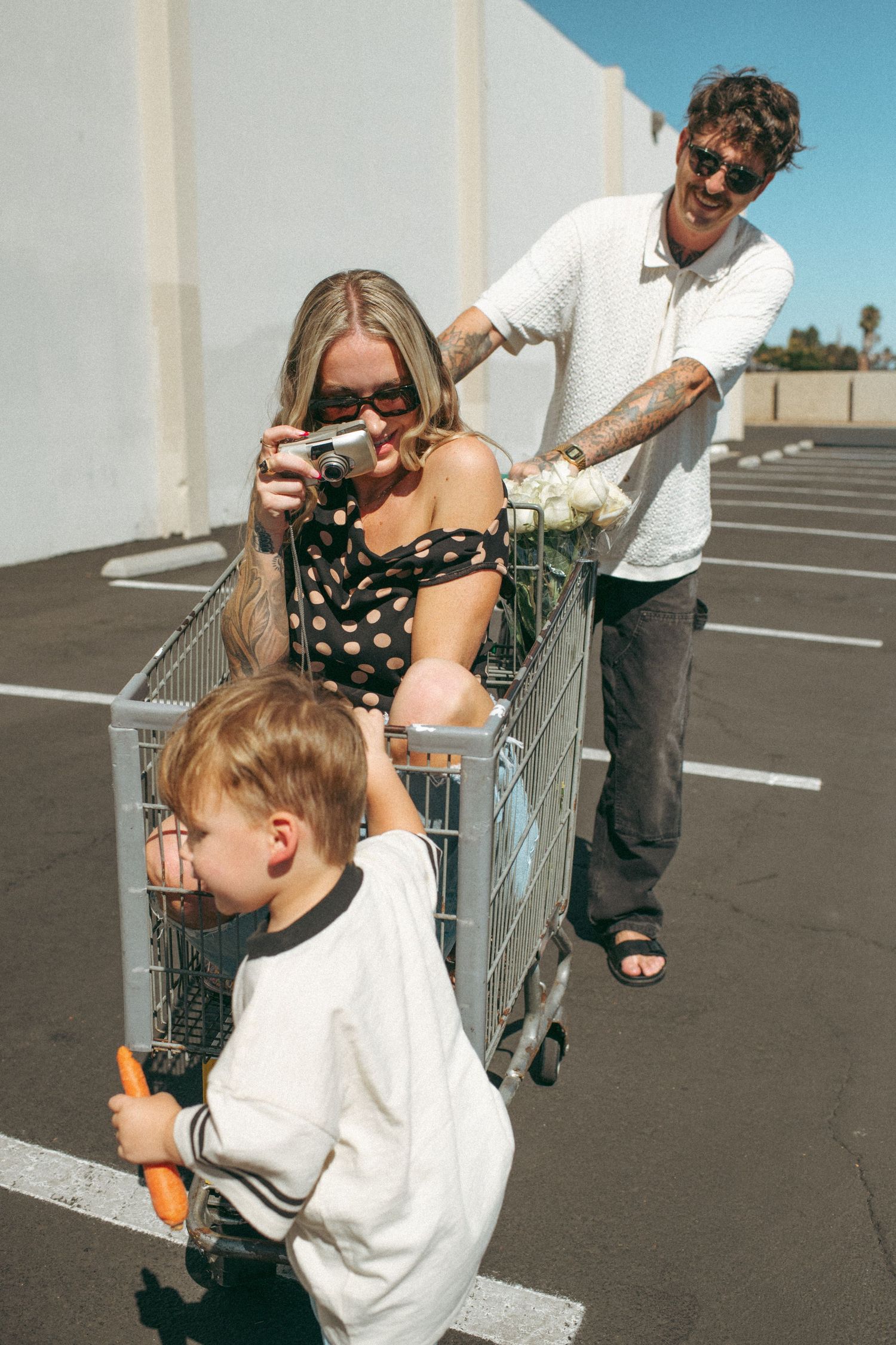Woman sitting in shopping cart taking photo while man holds flowers and toddler pushes cart in sunny parking lot.