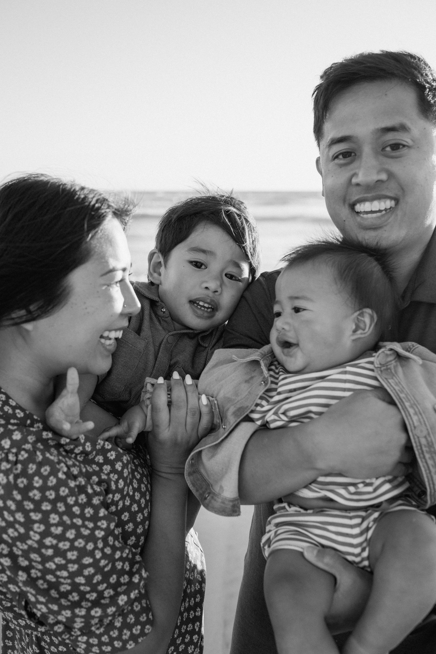 Black and white family portrait of parents with young son and baby at the beach, smiling and laughing together.