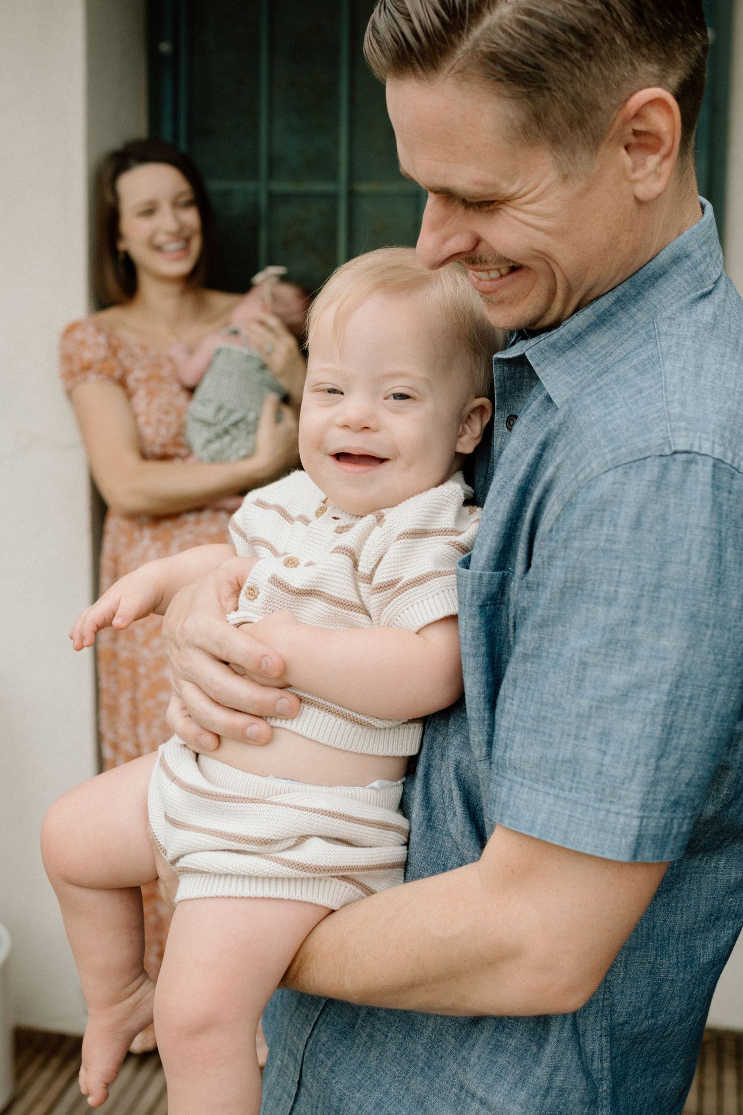 A smiling father holds a happy baby with Down syndrome while a mother stands behind holding a newborn.