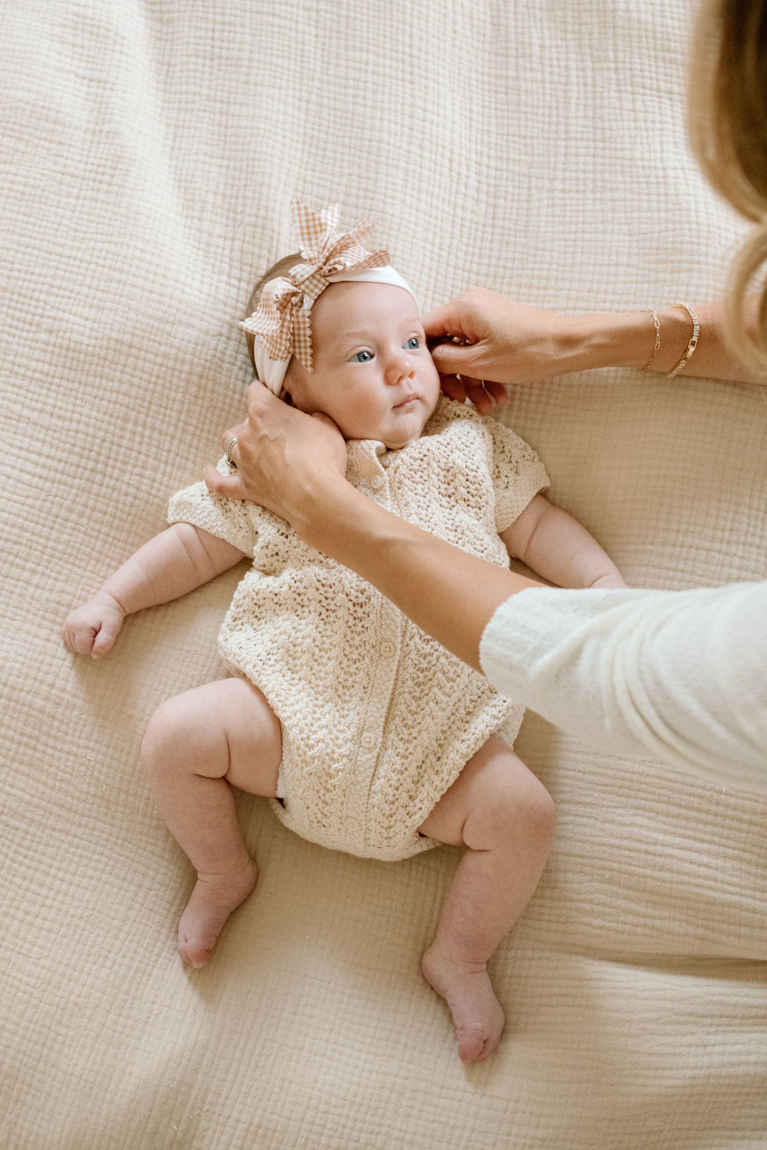 A mother adjusts a bow headband on her newborn baby girl in a cream knit romper lying on a beige blanket.