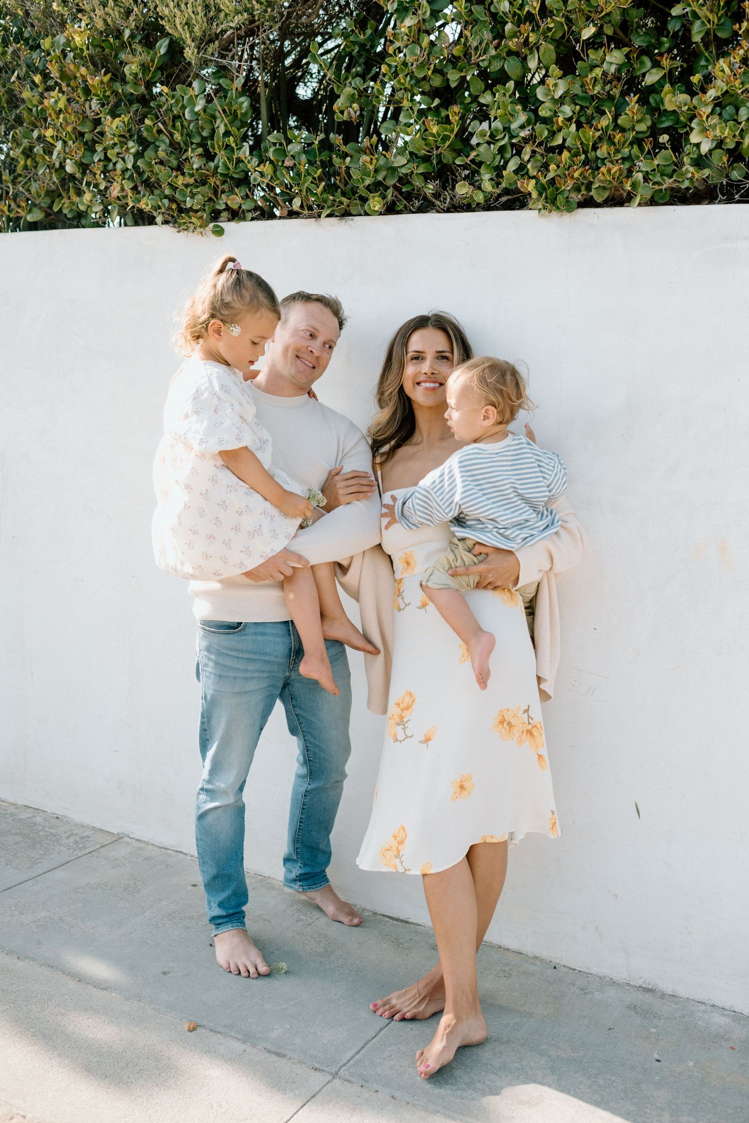 Happy family of four posing against a white wall, parents holding two young children, dressed in casual summer outfits.