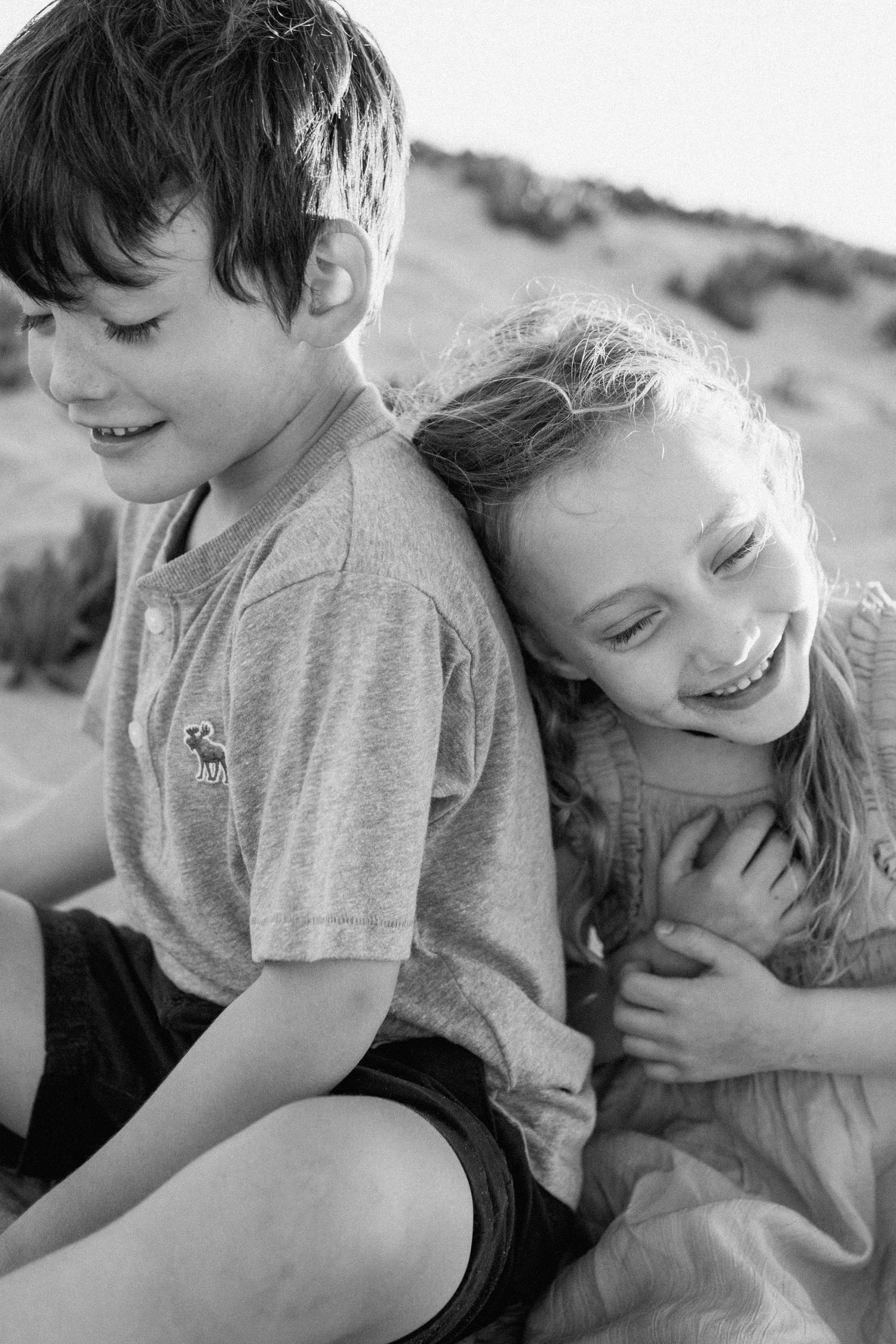 Black and white portrait of a laughing boy and girl sitting together on a beach.