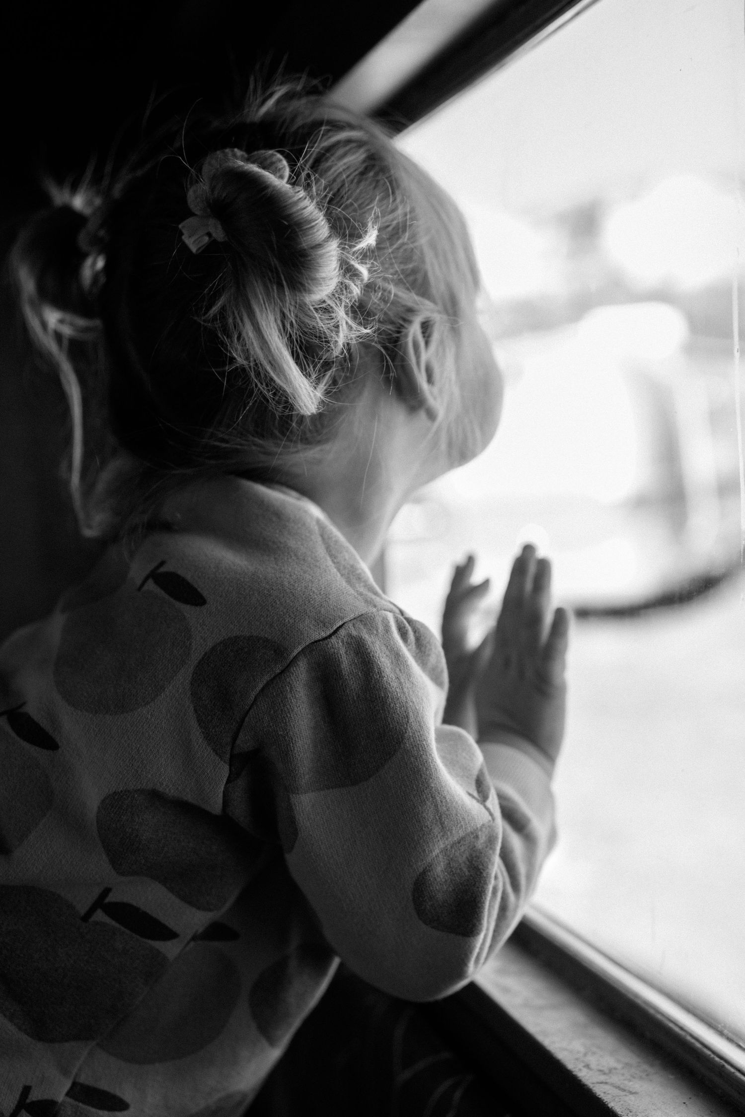 A black and white photo of a toddler girl looking out a window with hands pressed against the glass.