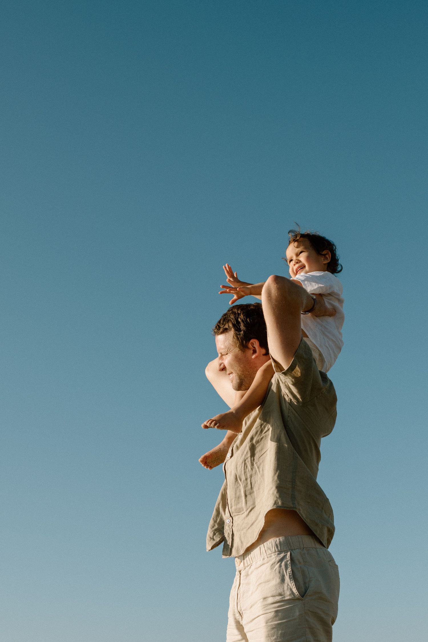 Father carrying young child on shoulders against clear blue sky, child spreading arms wide.