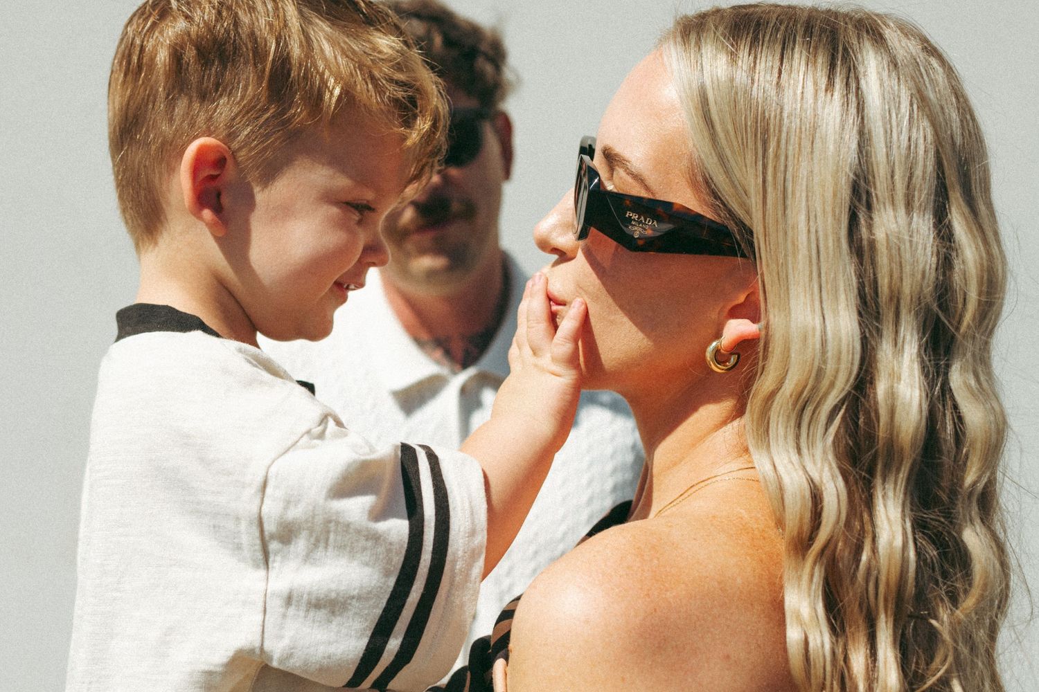 A young boy touches a blonde woman's face as she smiles in sunglasses, with a man visible in the background.