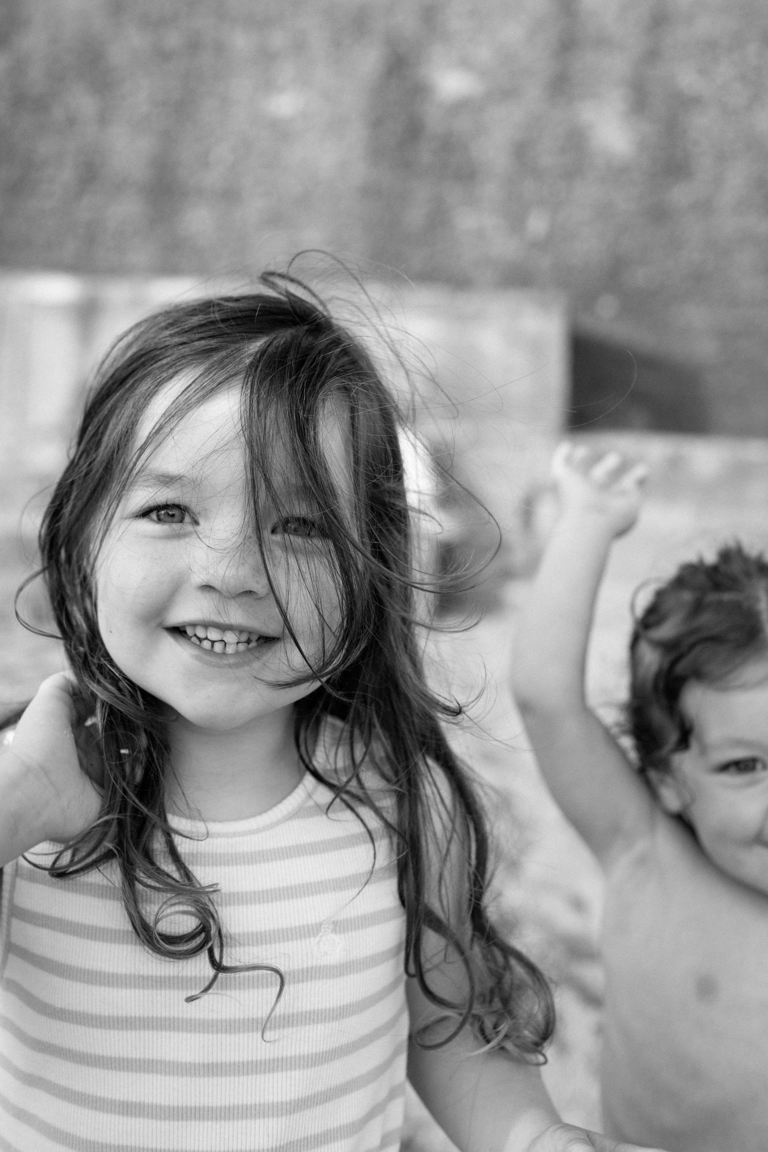 Black and white photo of a smiling young girl with windswept hair in a striped top, with a child in the background.