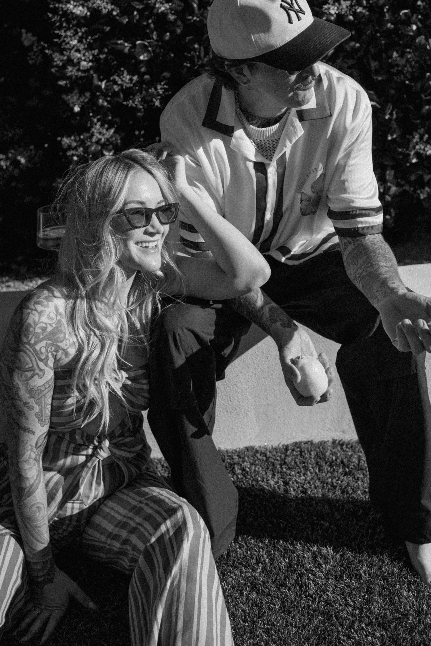 A black and white photo showing two people outdoors, with one wearing a Yankees cap and another in plaid pants sitting.