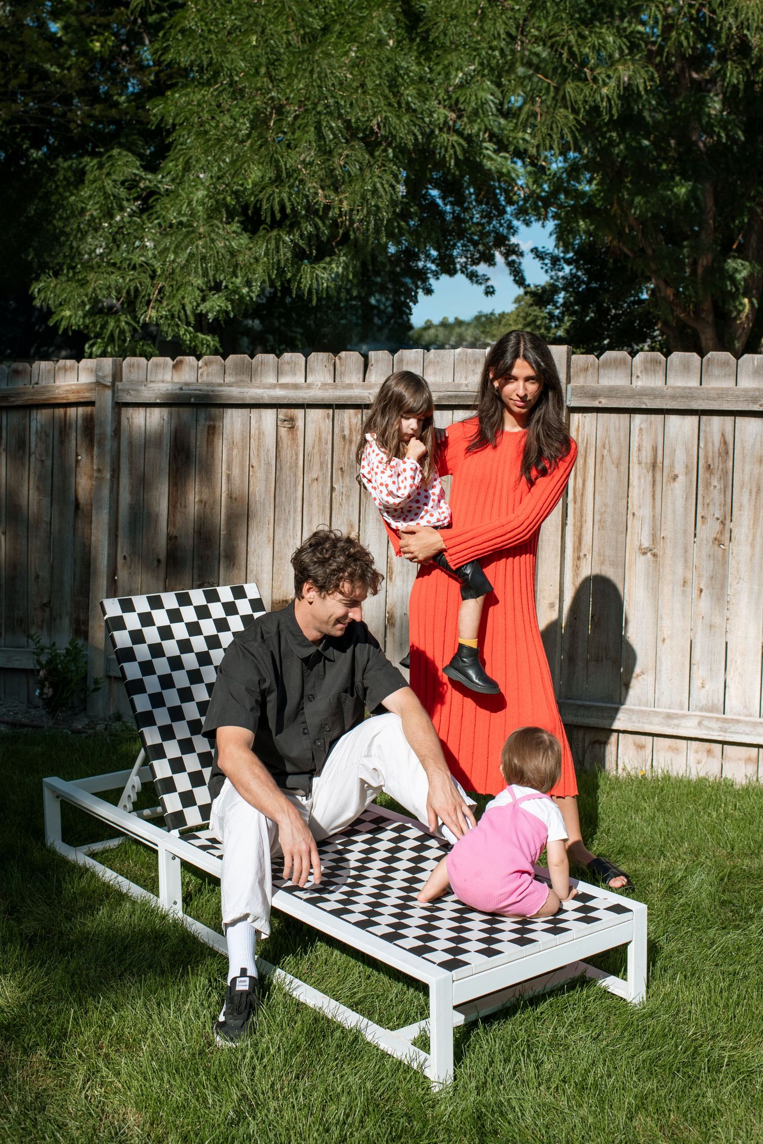Family of four relaxing outdoors on a checkered lounge chair in a sunny backyard with green grass and wooden fence.