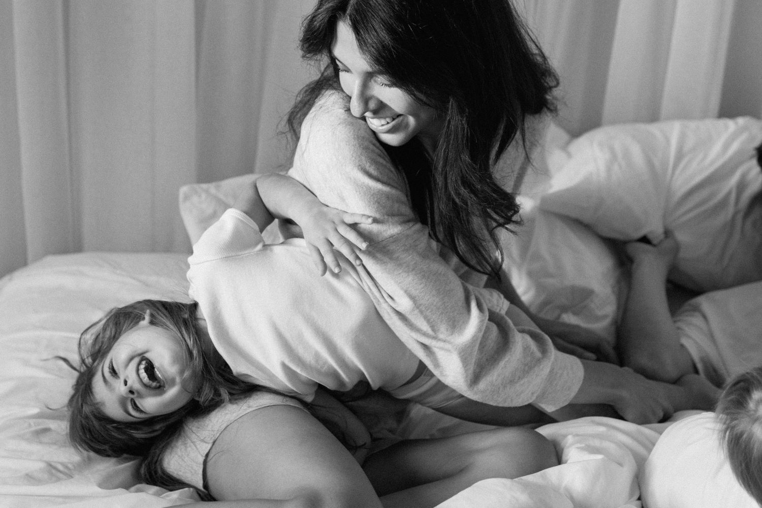 Black and white photo of a mother and daughter laughing and hugging playfully on a white bed.