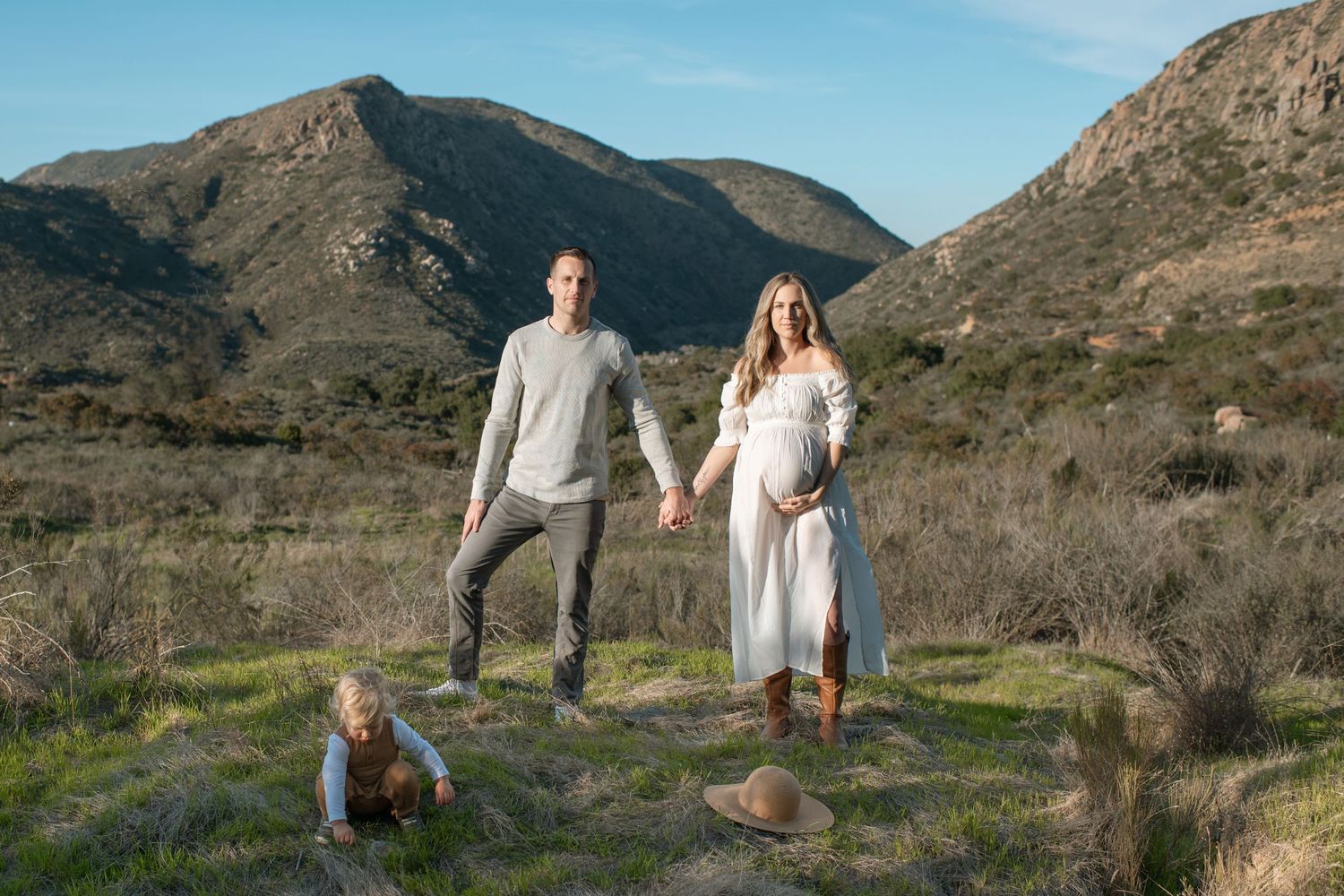 Pregnant woman in white dress with family posing in front of scenic mountain landscape outdoors.
