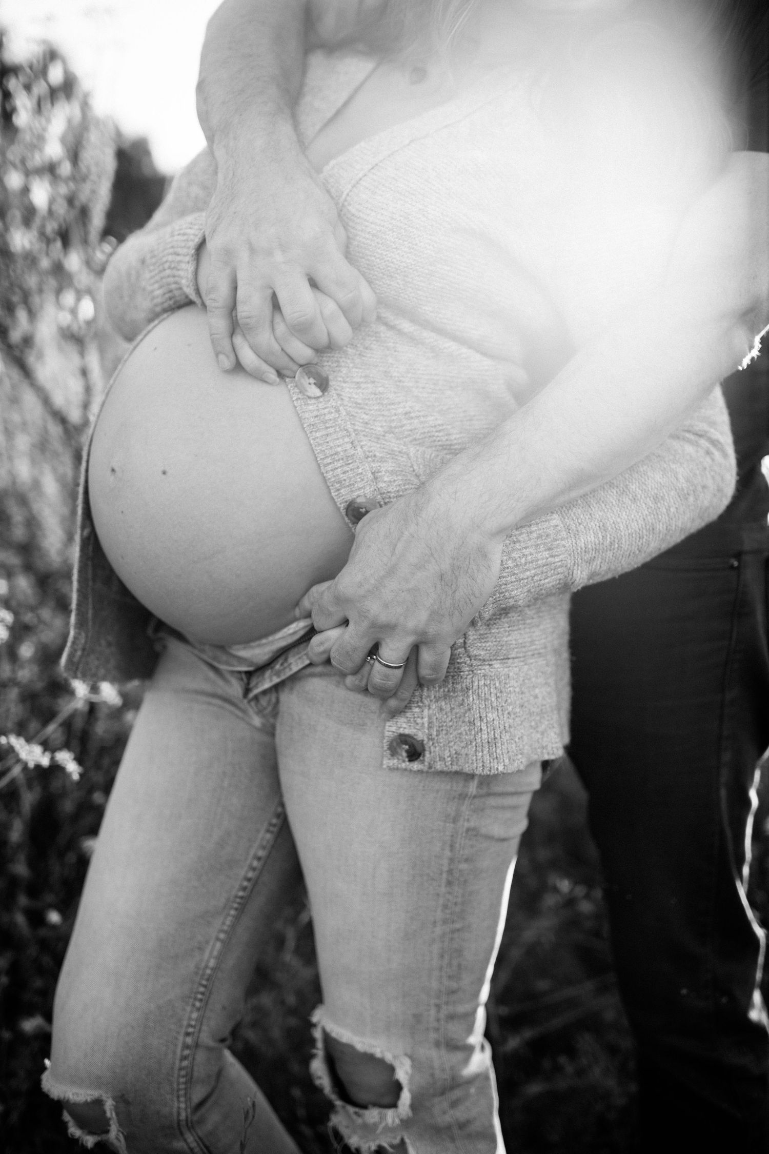 Black and white close-up of couple cradling pregnant belly outdoors in bright sunlight.