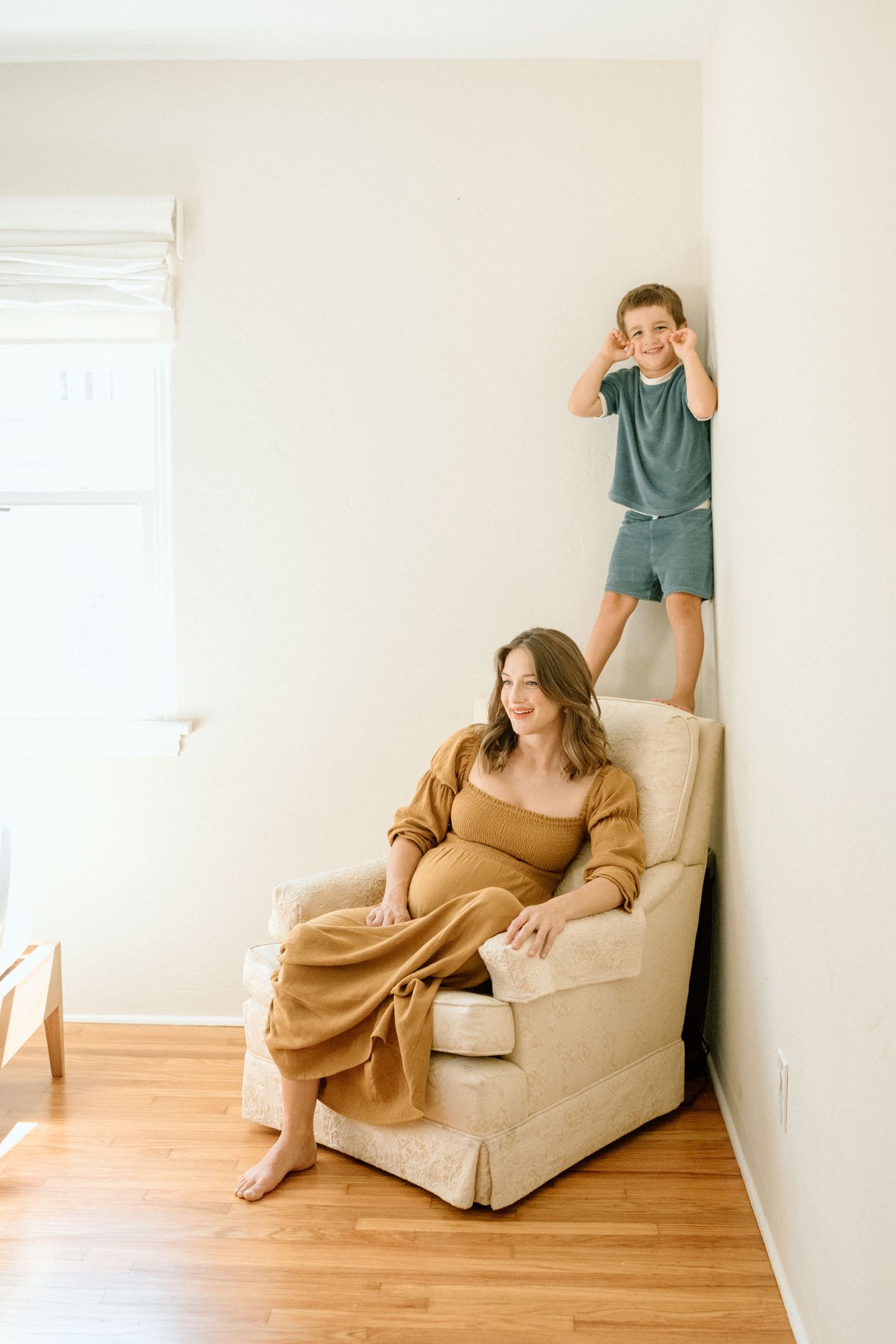 Pregnant woman sitting in armchair wearing mustard outfit while young boy stands on chair arm behind her.