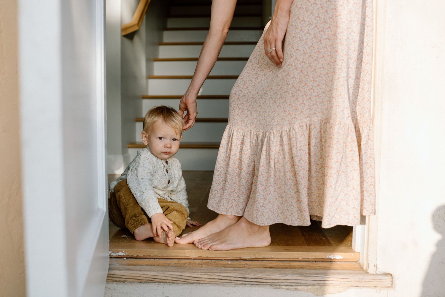 Baby sitting on staircase landing beside mother in floral dress holding his hand.