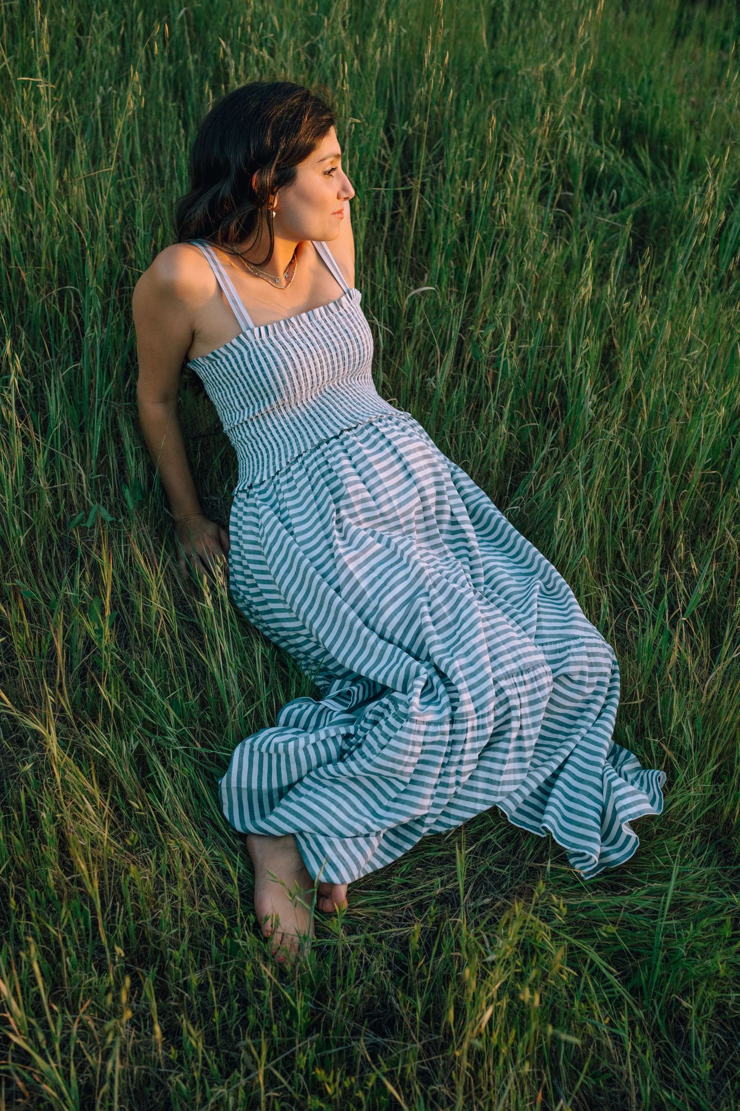 Pregnant woman in striped sundress sitting in tall green grass at sunset.