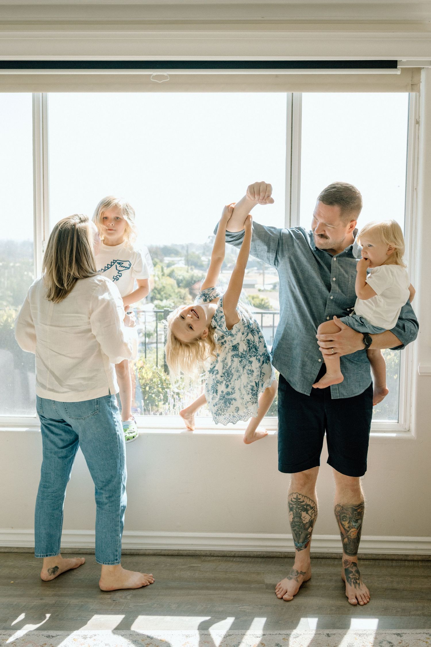 A joyful family playing together indoors, with tattooed dad swinging a child while mom holds another near a bright window.