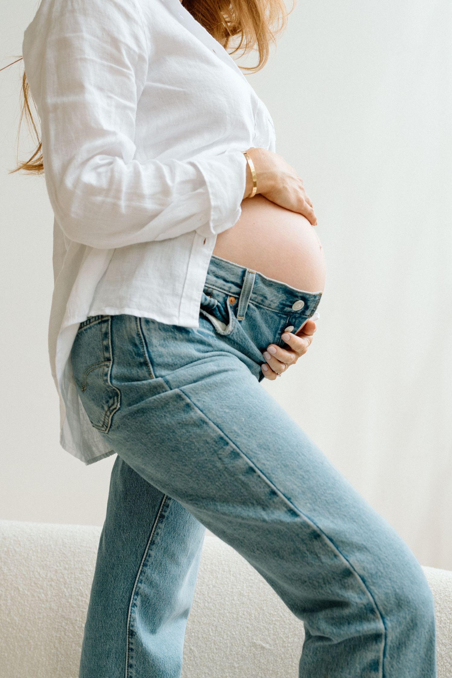 Pregnant woman in white linen shirt and jeans cradling her bare baby bump.