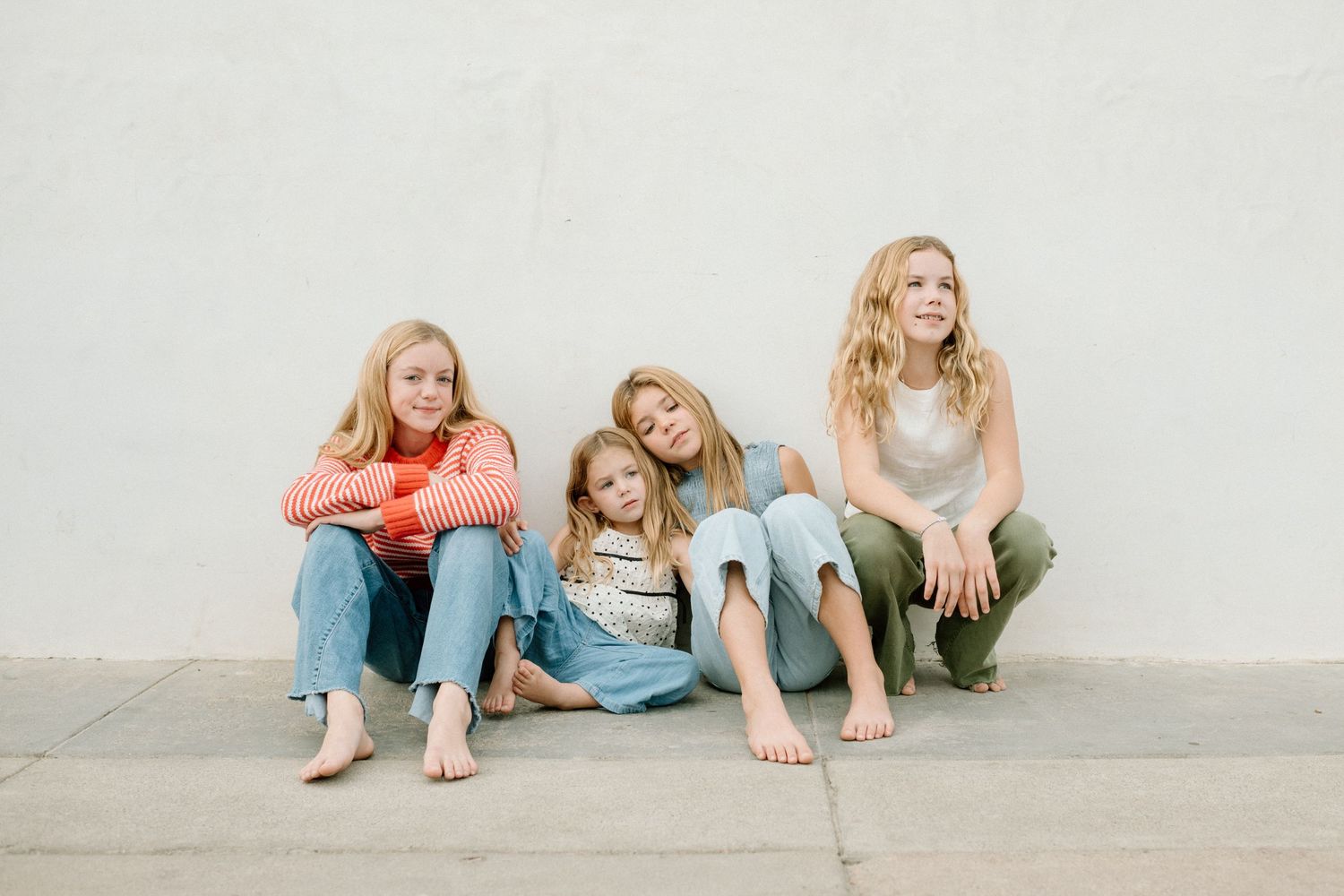 Four barefoot blonde girls sitting and leaning against a white wall, smiling and looking upward casually.