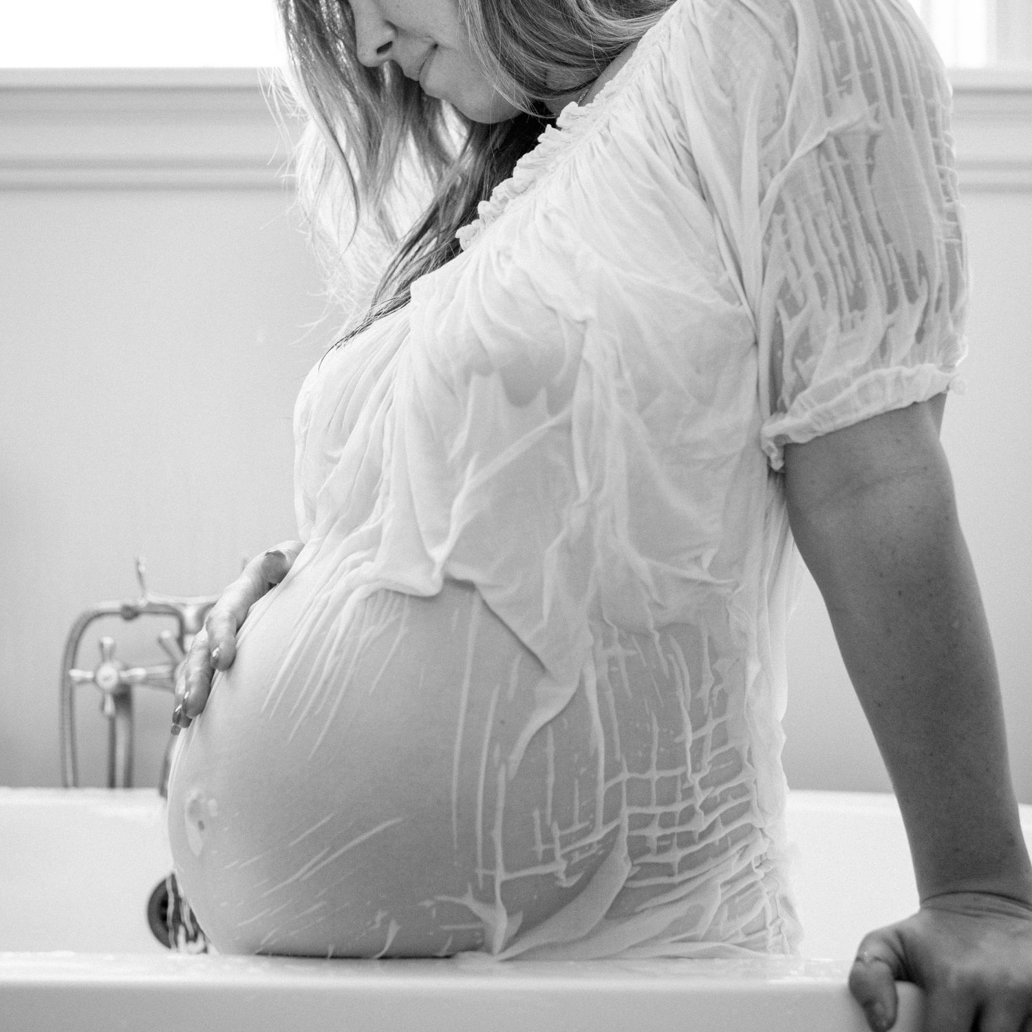 Pregnant woman in wet white sheer top leans over bathtub cradling her baby bump in black and white photo.
