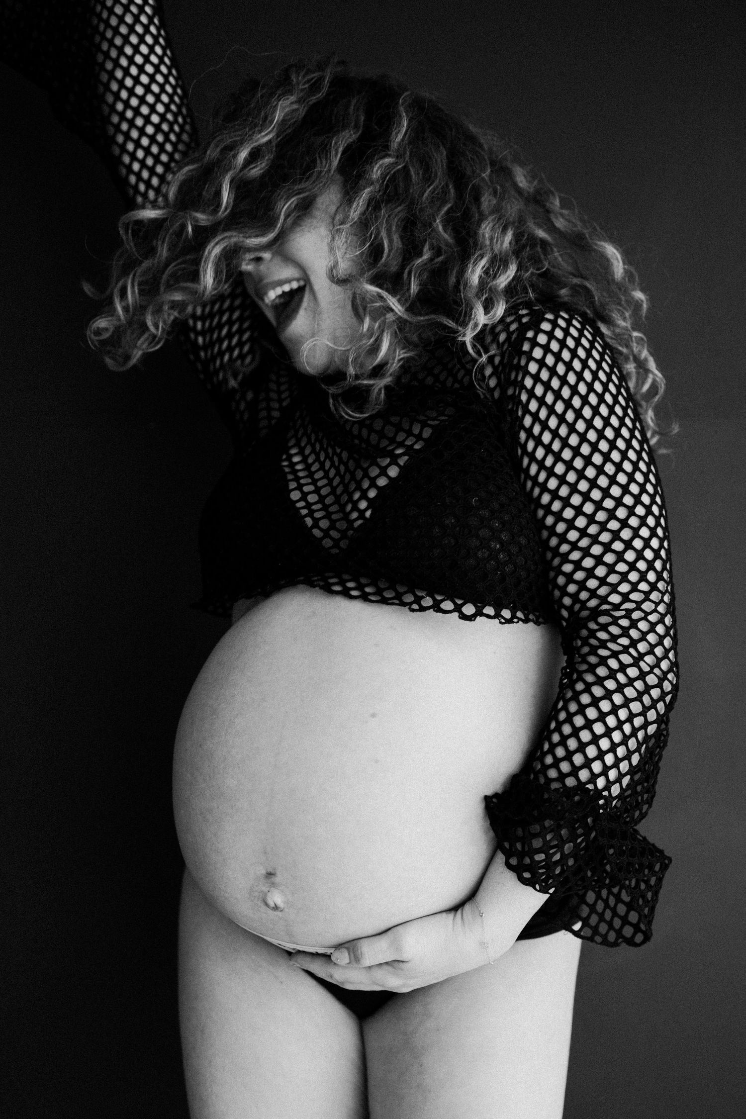 Joyful pregnant woman in fishnet top holding belly in black and white maternity photo.