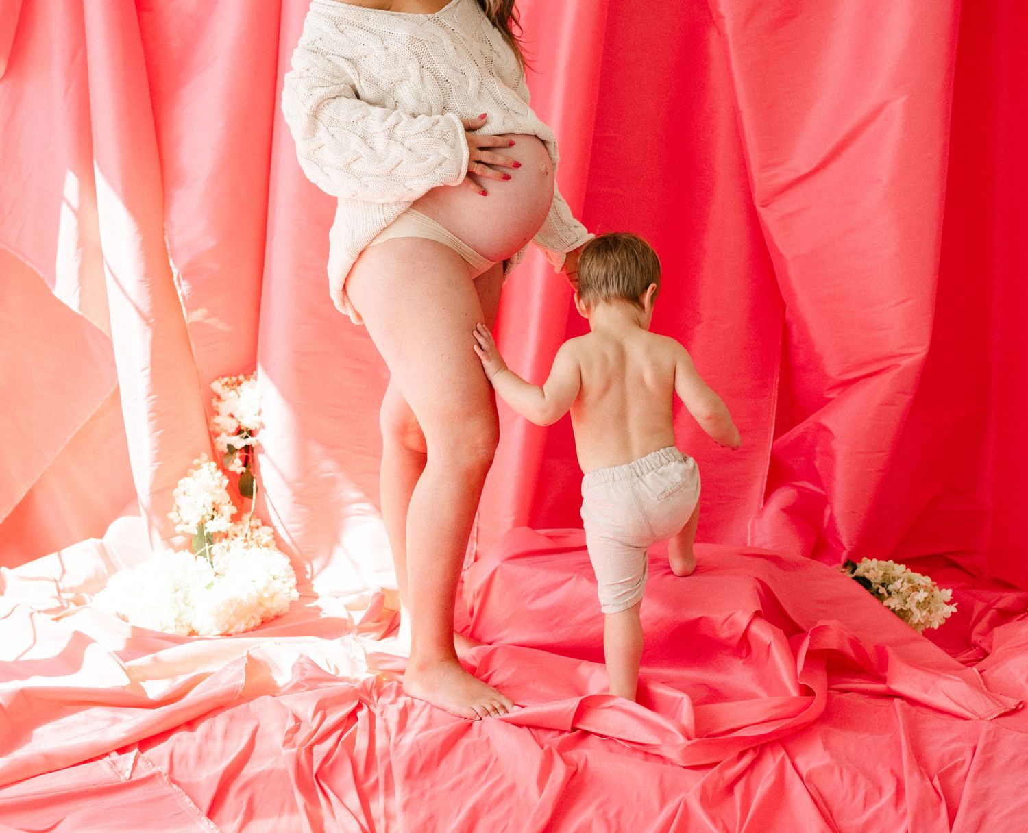 Pregnant woman in white top with toddler in diaper standing on pink fabric backdrop in maternity photo shoot.