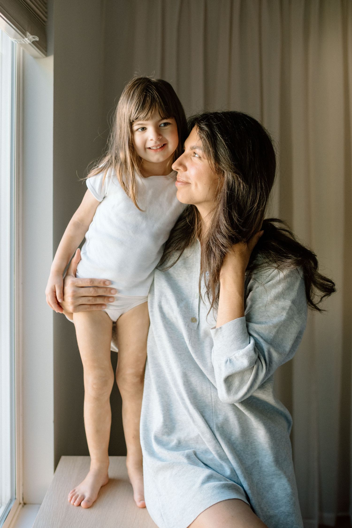 A mother holds her young daughter near a window, both smiling in casual home attire.