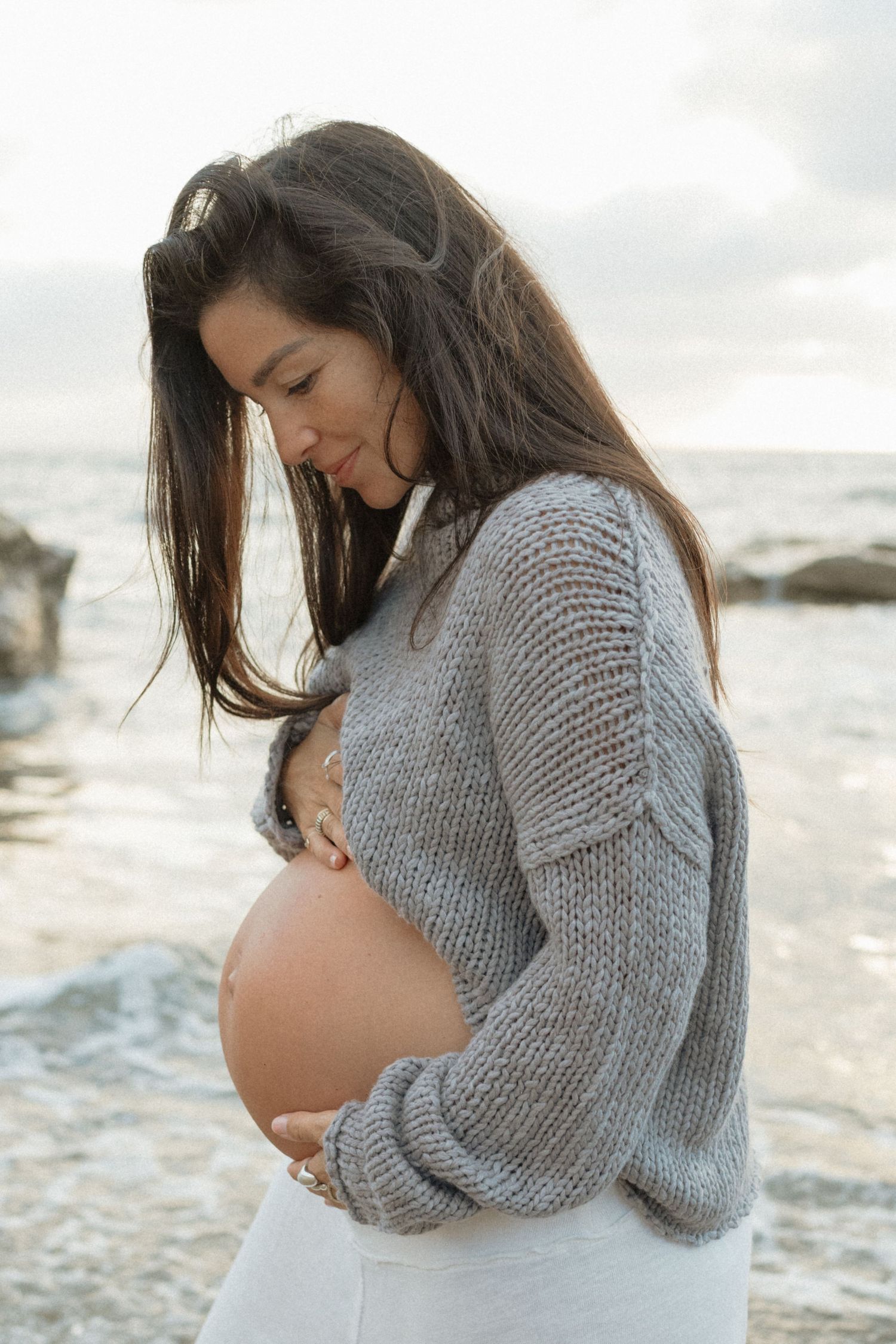 A pregnant woman in a gray sweater cradles her baby bump while standing near the ocean shore.