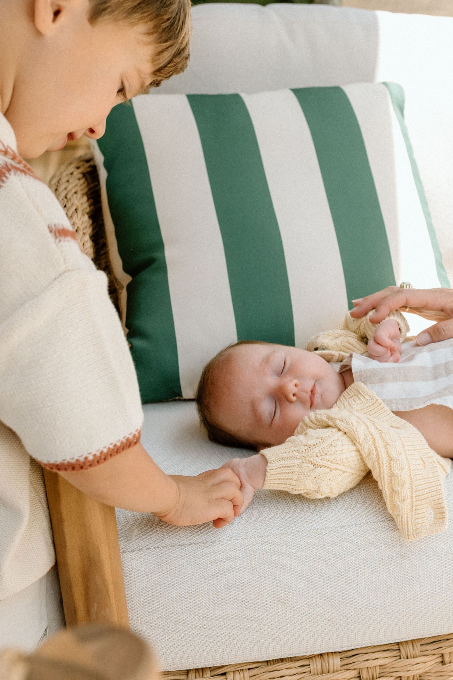 Young boy gently holding sleeping newborn baby's hand on striped outdoor chair.