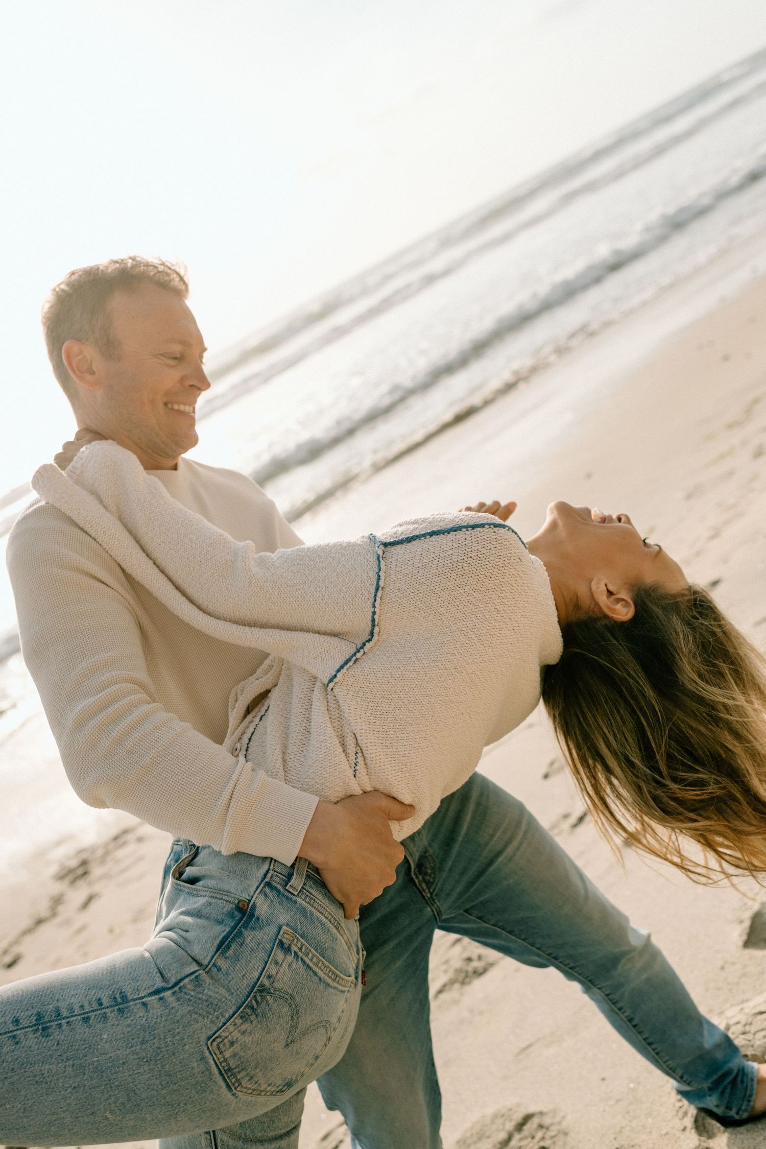Happy couple laughing and dancing together on a sunny beach, wearing casual jeans and sweaters.