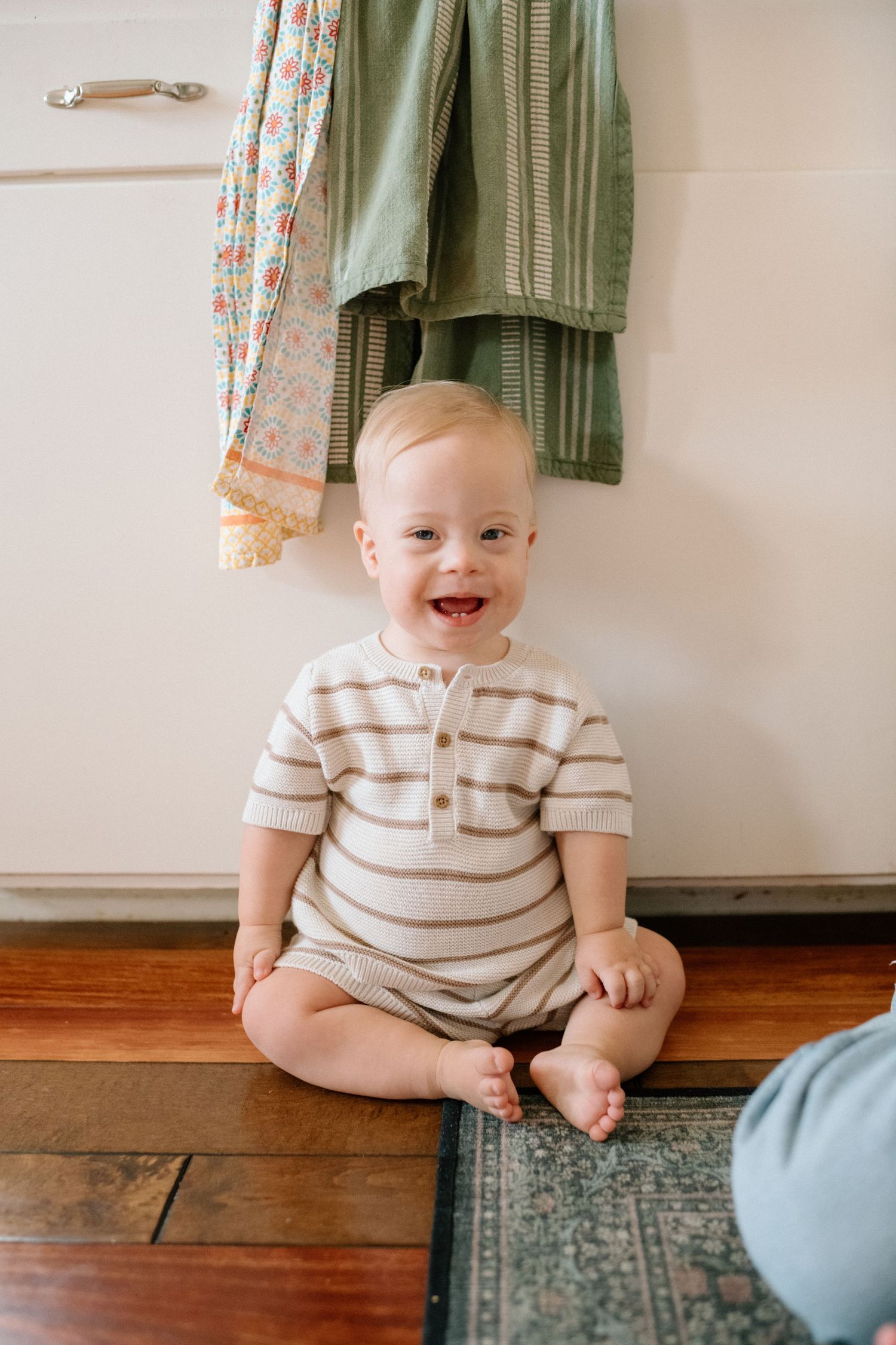 Happy baby in striped onesie sitting on hardwood floor smiling at camera.